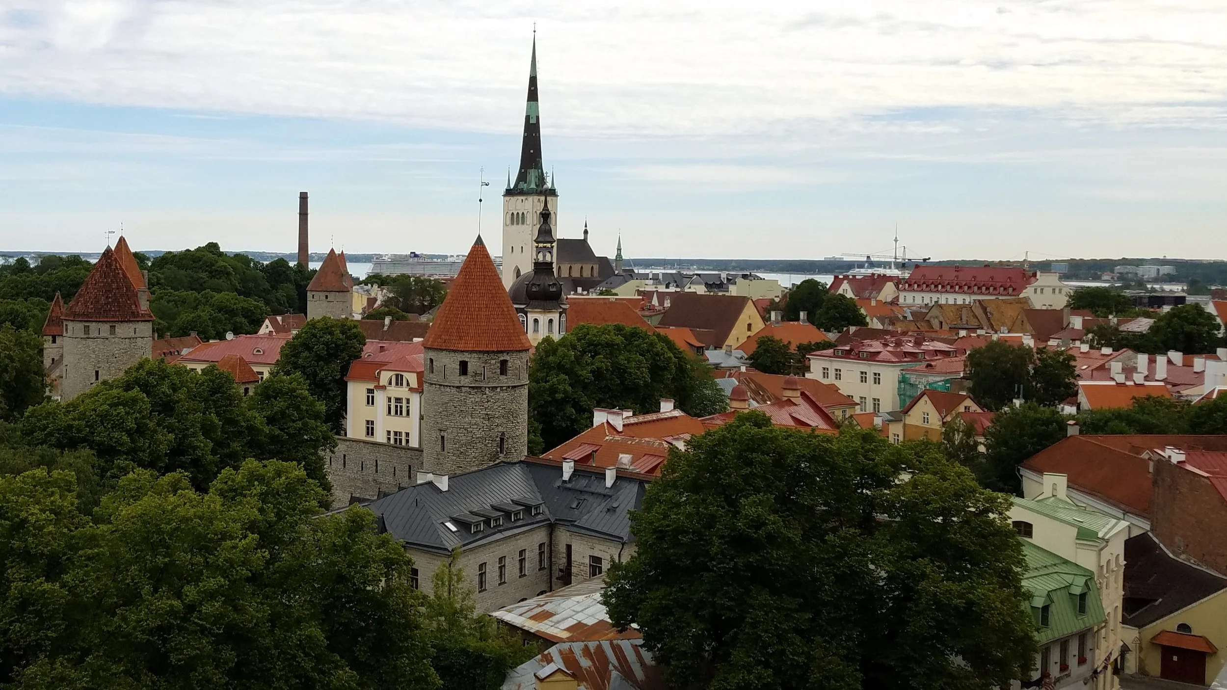 A panoramic view of Tallinn, Estonia, featuring historic buildings with red and gray tiled roofs, several church spires, towers, lush green trees, and a distant harbor with ships and wind turbines under a cloudy sky.