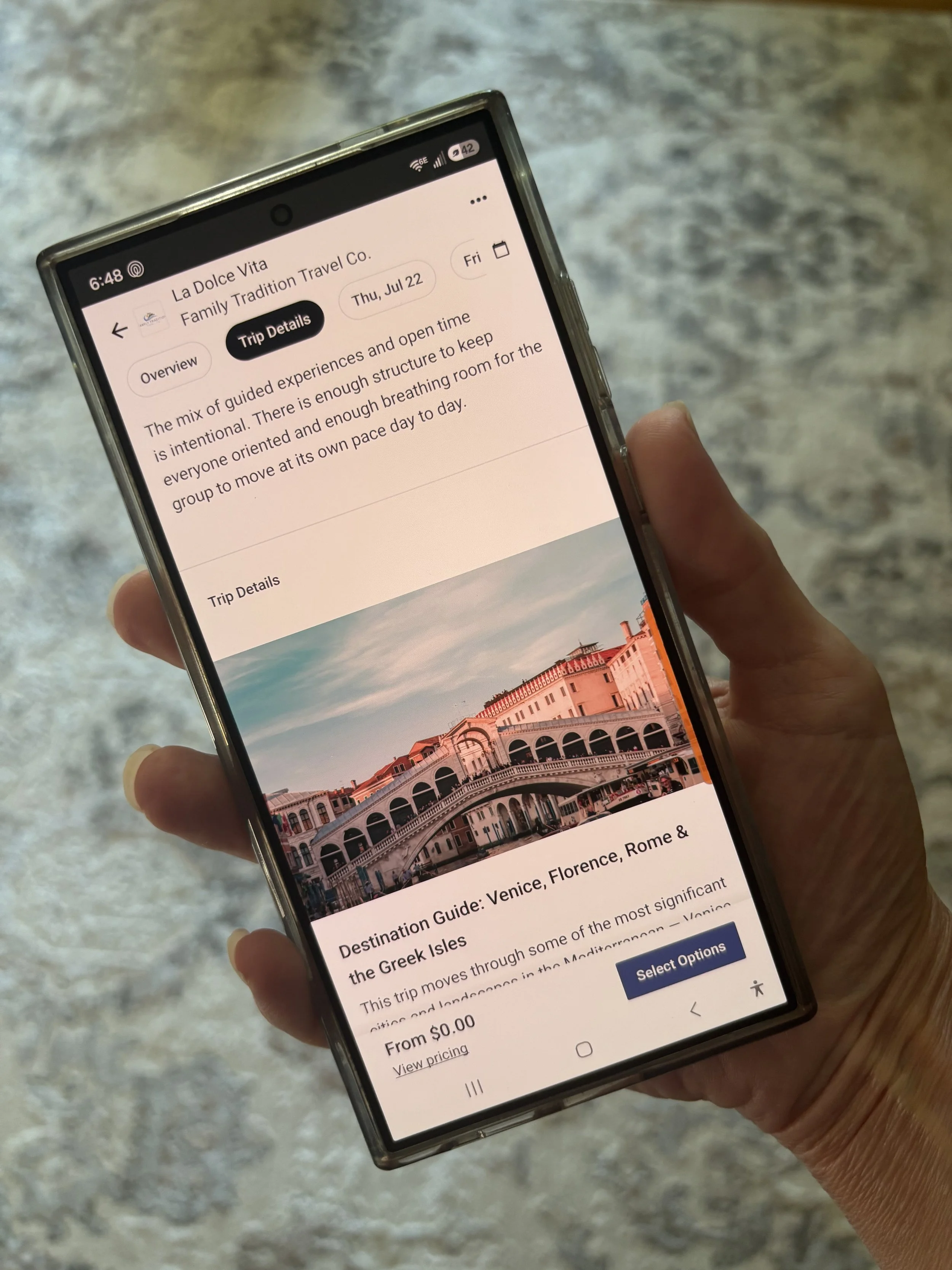 A person holding a smartphone displaying a travel app with information about a trip to Venice, Florence, Rome, and the Greek Isles, with a photo of a Venice canal and buildings.