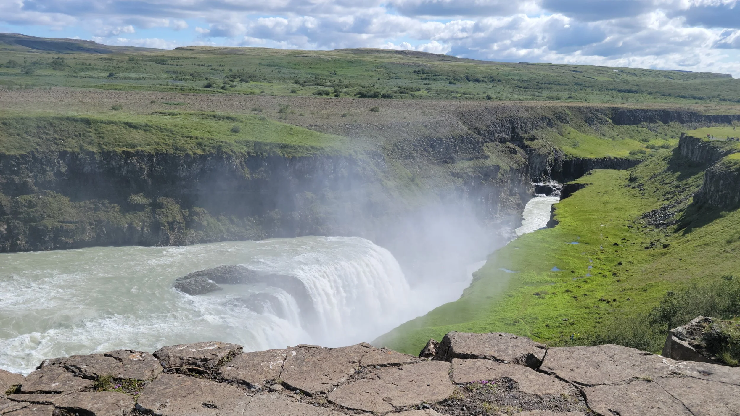 A wide view of a powerful waterfall flowing through a lush green valley with cliffs on either side, under a partly cloudy sky.