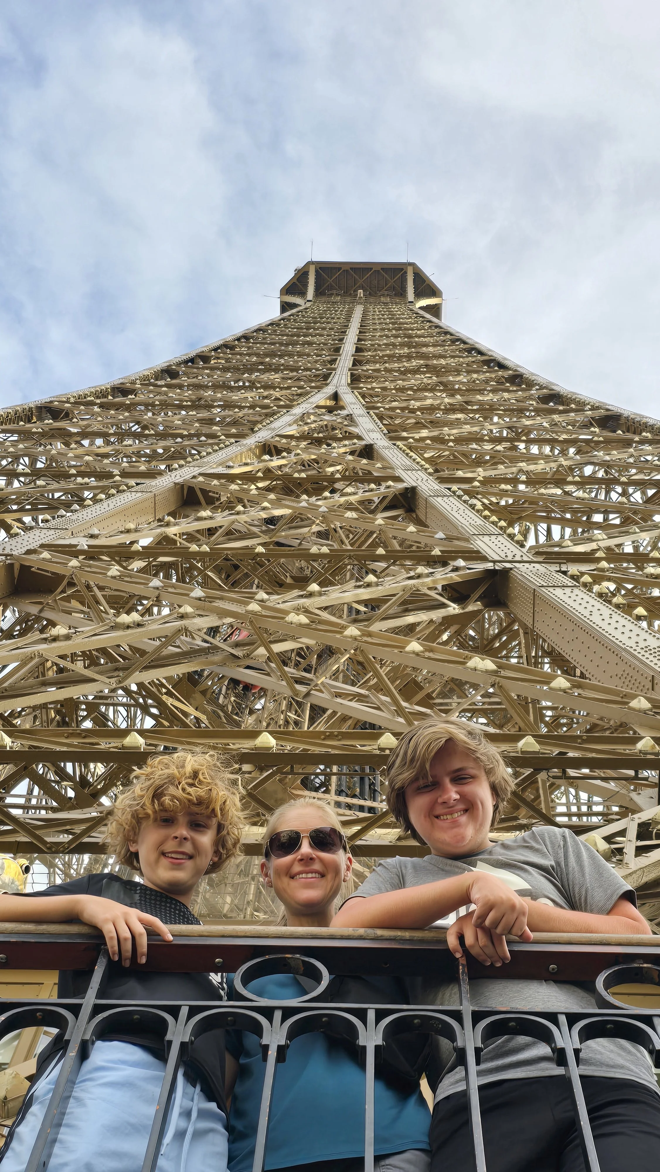 A mother and her two teenage sons pose at the railing of the Eiffel Tower's lower platform, with the tower's iron structure rising above them.