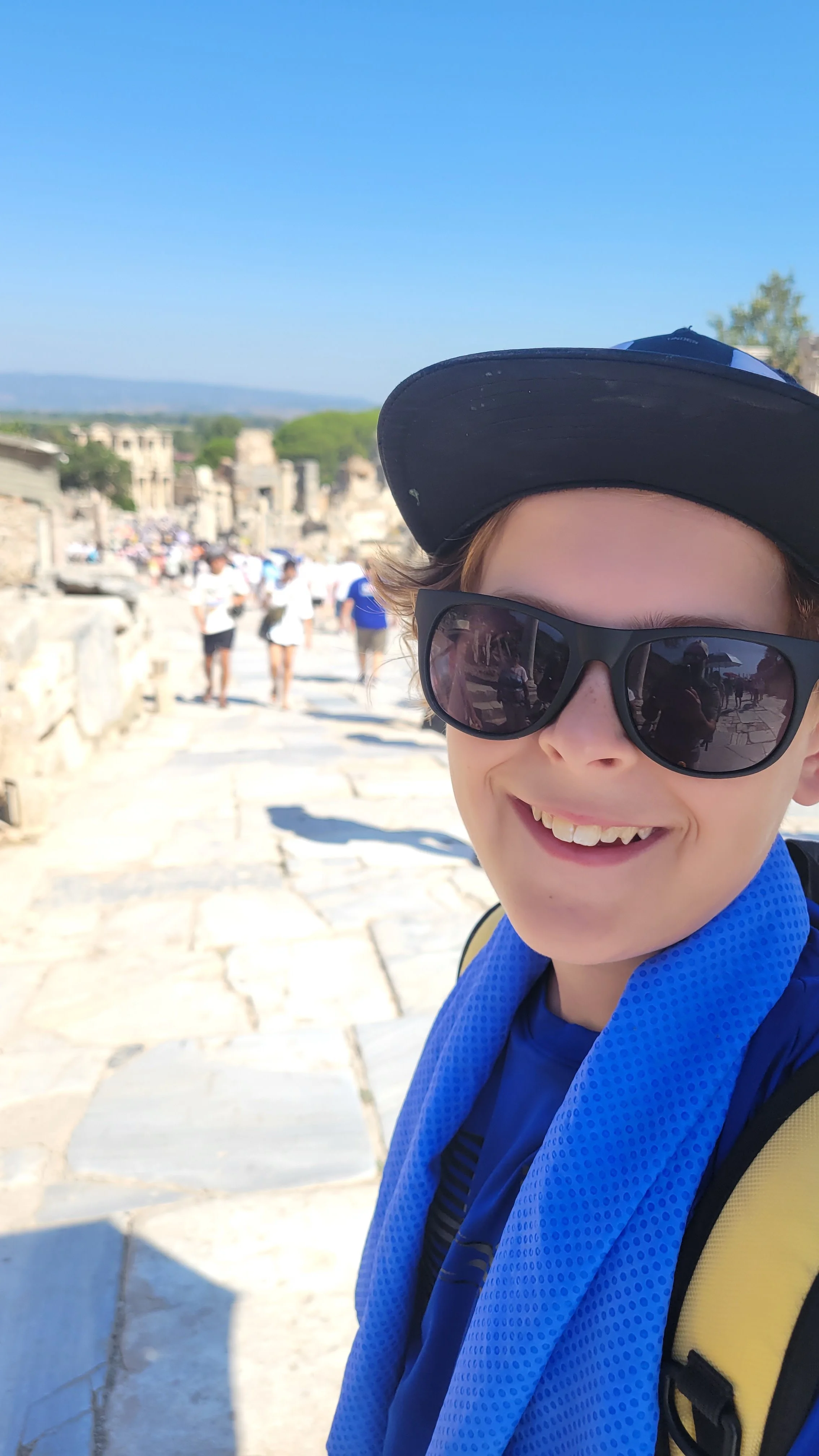 A boy wearing sunglasses and a blue cooling towel around his neck on the ancient stone streets of Ephesus, Turkey, with visitors and ruins in the background.