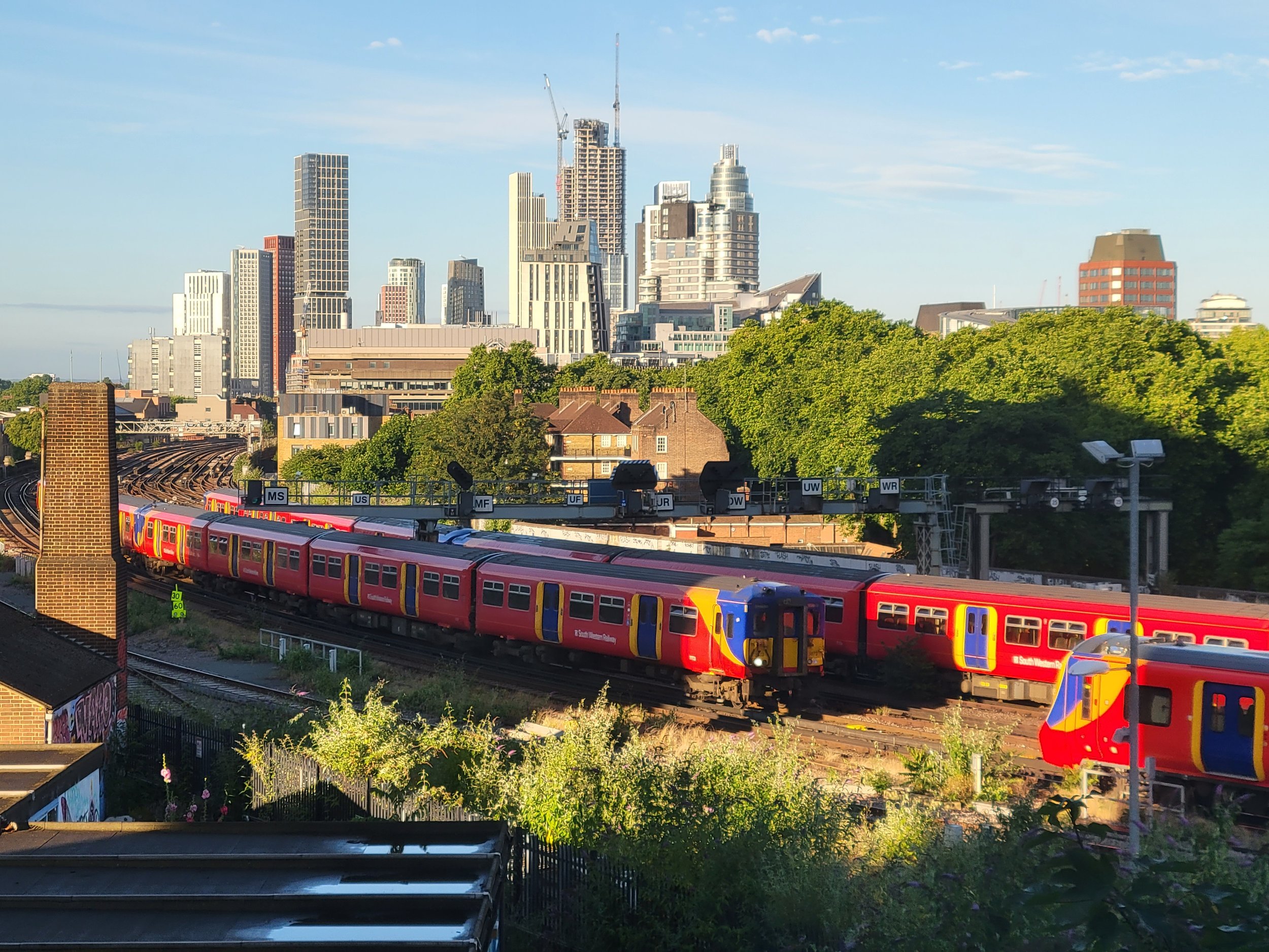 View of a city skyline with tall buildings, some under construction, and a train moving on elevated tracks in the foreground. Green trees are visible in the lower part of the image, and the sky is mostly clear with some scattered clouds.