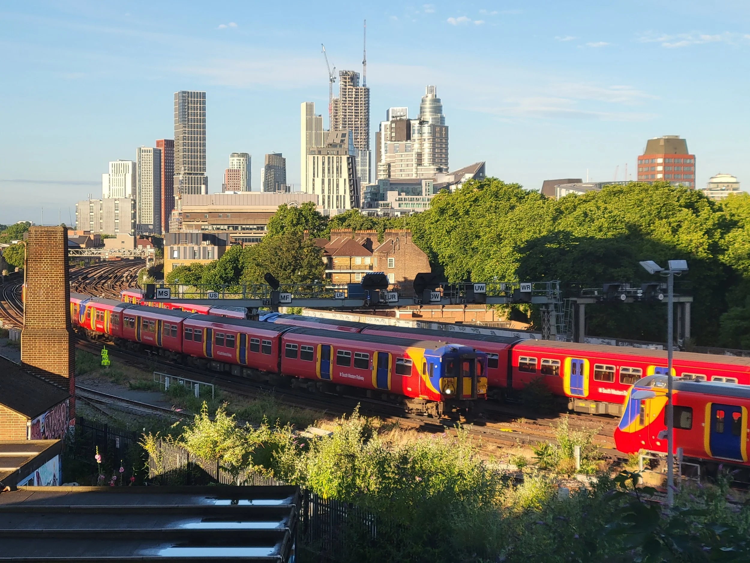 View of a city skyline with tall buildings, some under construction, and a train moving on elevated tracks in the foreground. Green trees are visible in the lower part of the image, and the sky is mostly clear with some scattered clouds.