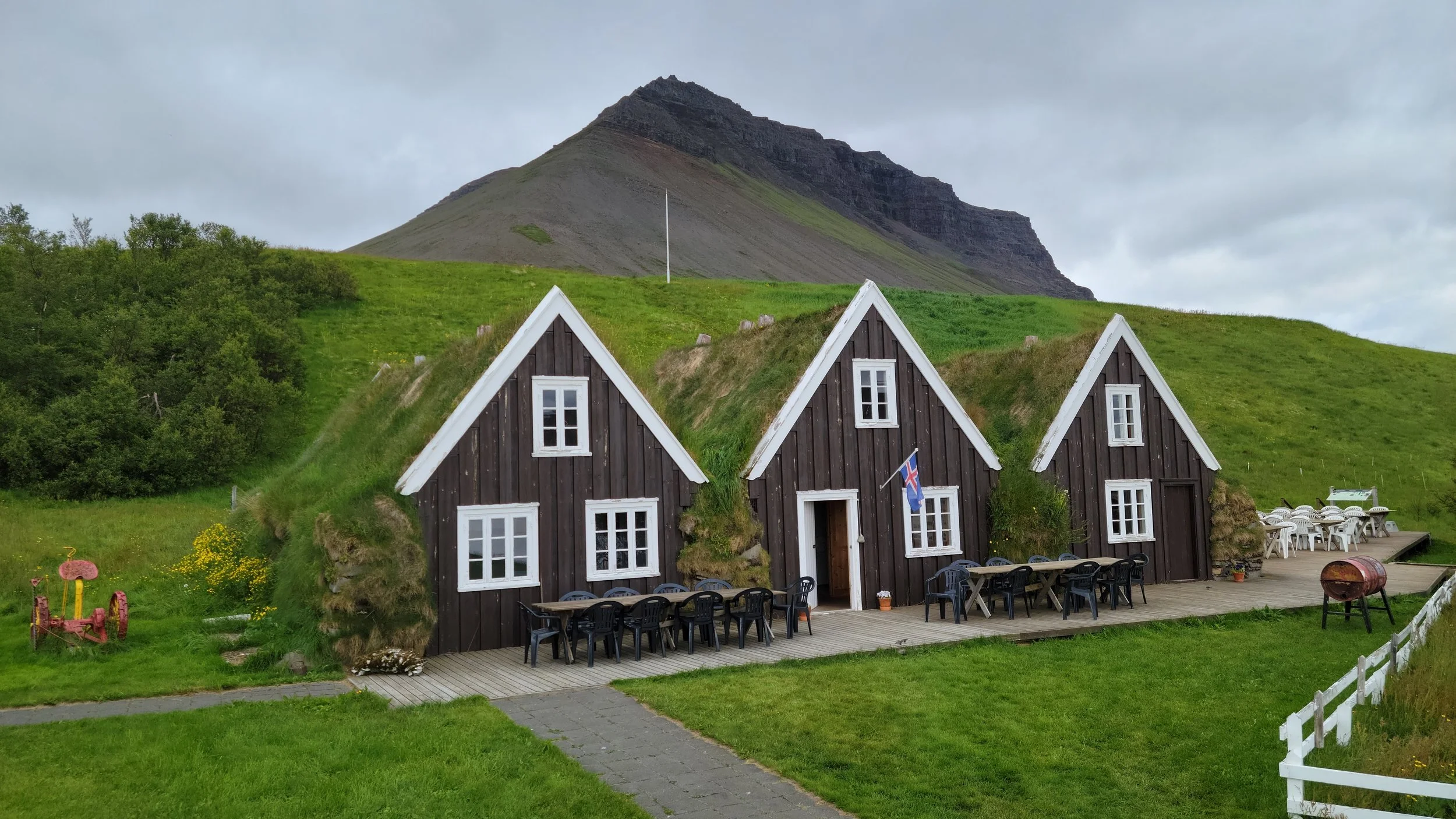 A small traditional Icelandic black wooden house with a grass roof, situated on a grassy hillside with a mountain in the background. There are outdoor tables and chairs outside, a flag on the house, and greenery surrounding the area.
