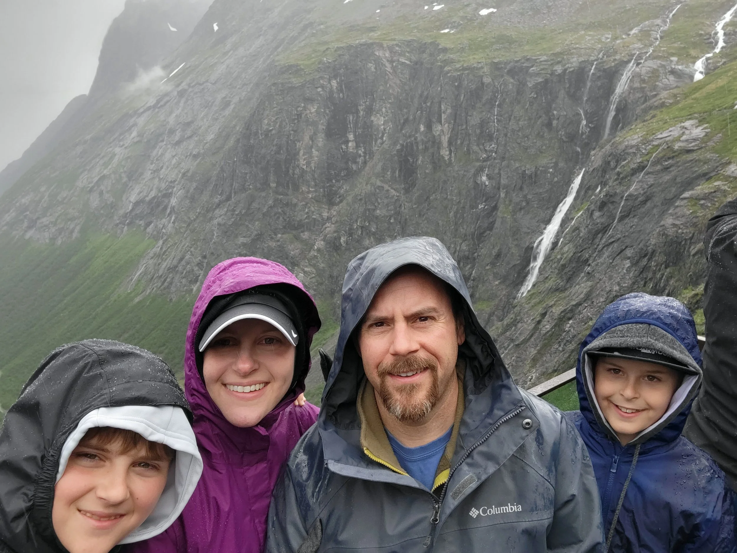 Family of four in rain jackets with hoods up, smiling in front of a misty cliff face with waterfalls, in Ålesund, Norway.
