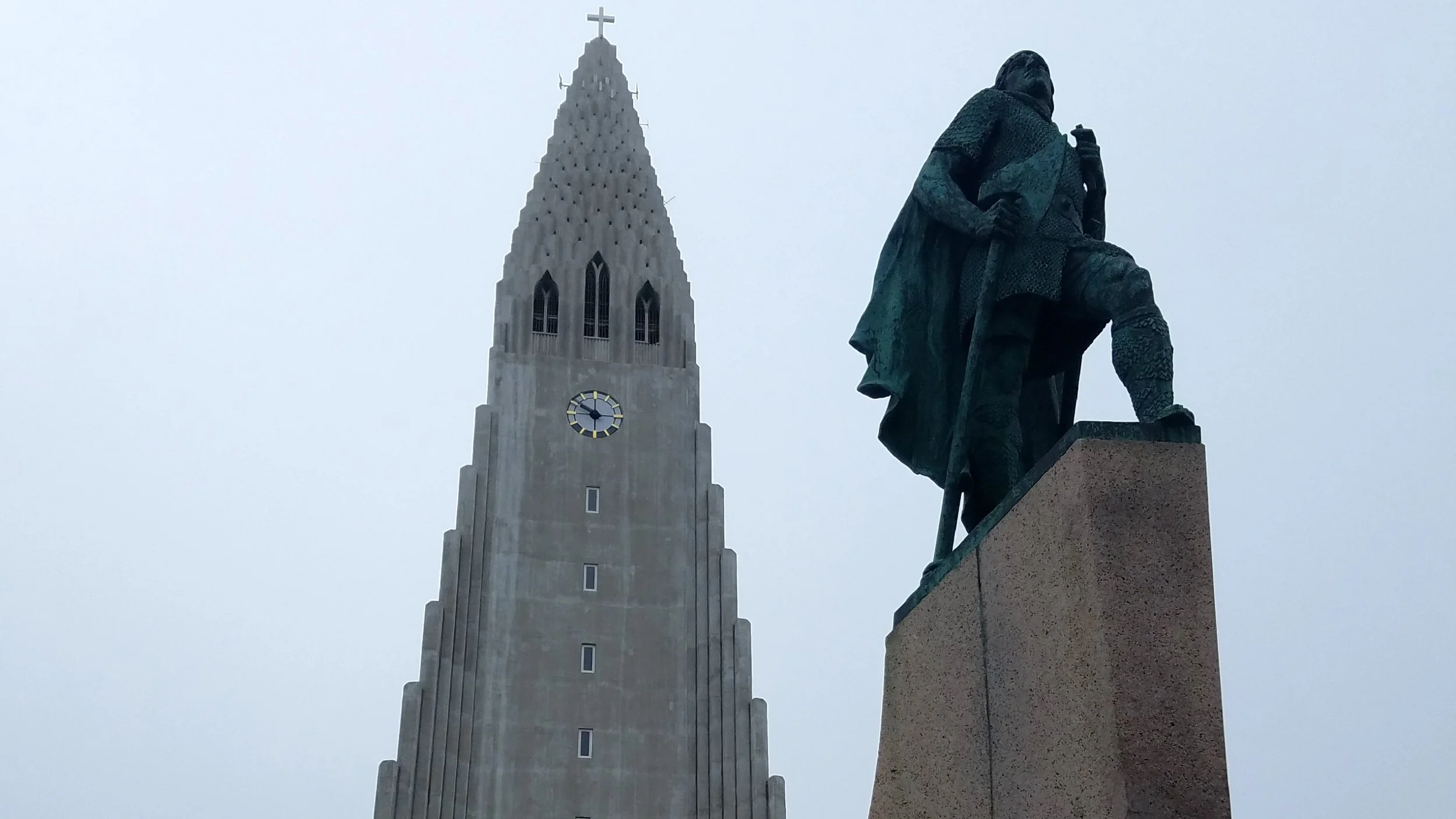 A statue of a seated person holding a staff, situated on a pedestal, with a tall church tower with a clock and a cross on top visible in the background on a cloudy day.