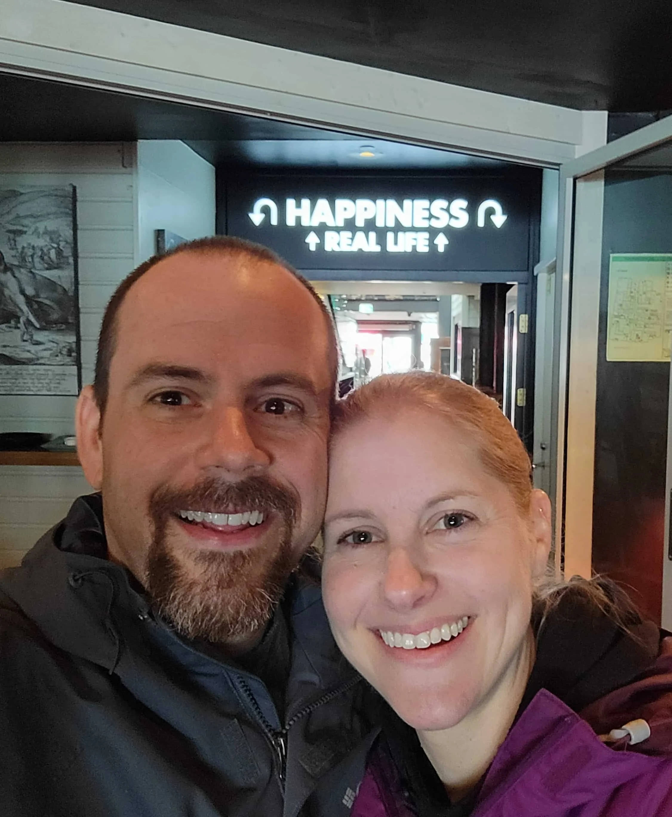 Two smiling people, a man with a beard and a woman with blonde hair, taking a selfie together. Behind them, a sign that reads 'Happiness = Real Life' with arrows pointing up and down.