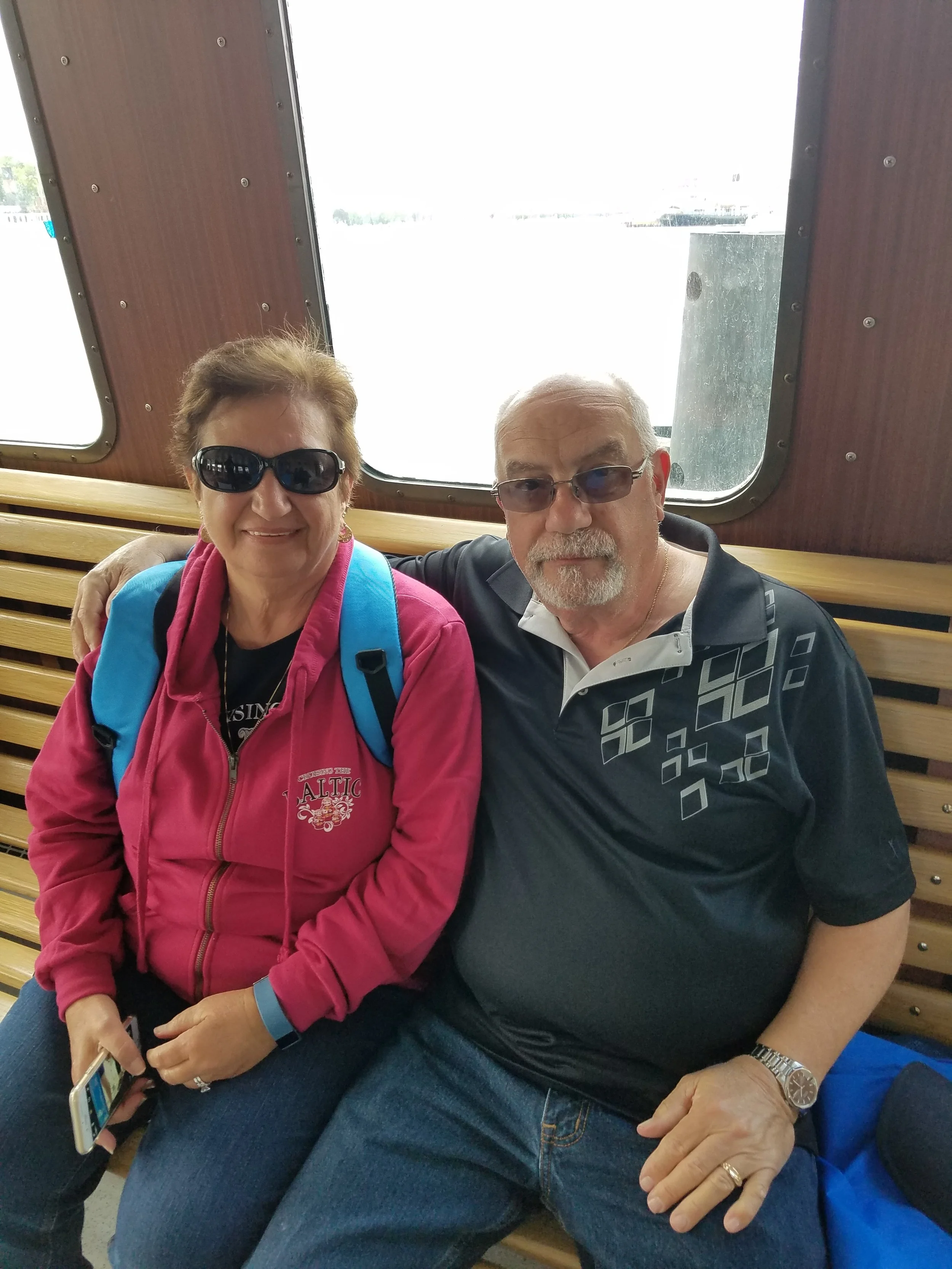 An older couple sits together on a wooden bench inside a passenger ferry in Stockholm, Sweden, with harbor water visible through the window behind them.