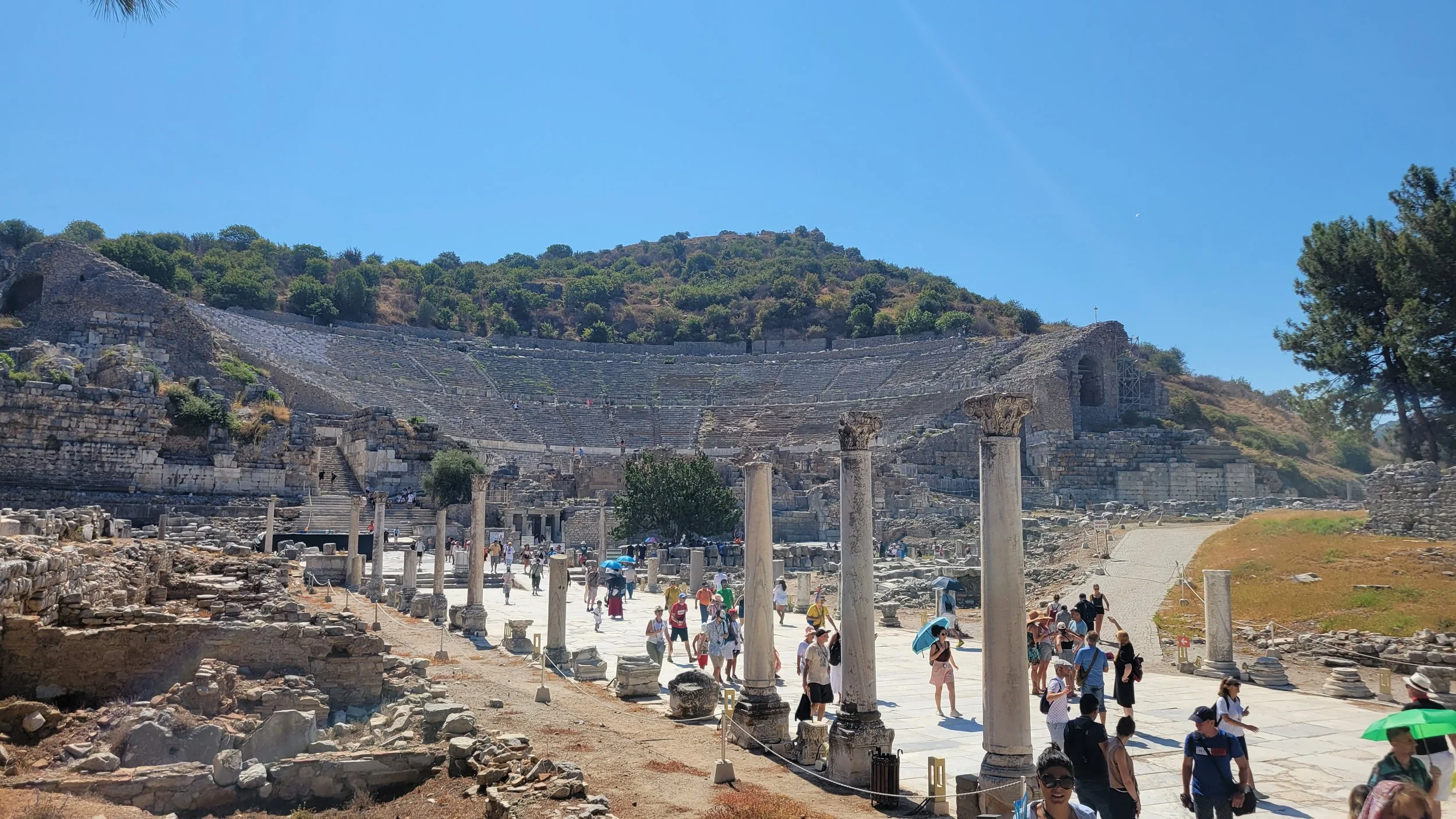 Ancient Greek amphitheater with stone columns and descending stairs, visitors walking around, hills with greenery in the background under a clear blue sky. Ephesus Archaeological Site, Türkiye