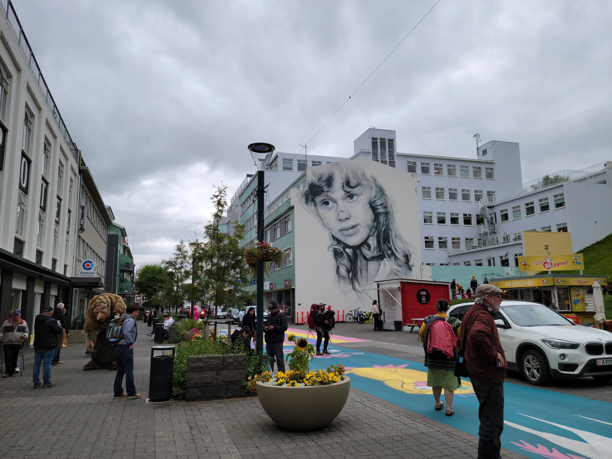 A busy city street with people walking and gathering. There is a large mural of a young girl on the building, and shops with signs. The sky is overcast, and there are flowers and planters along the sidewalk.