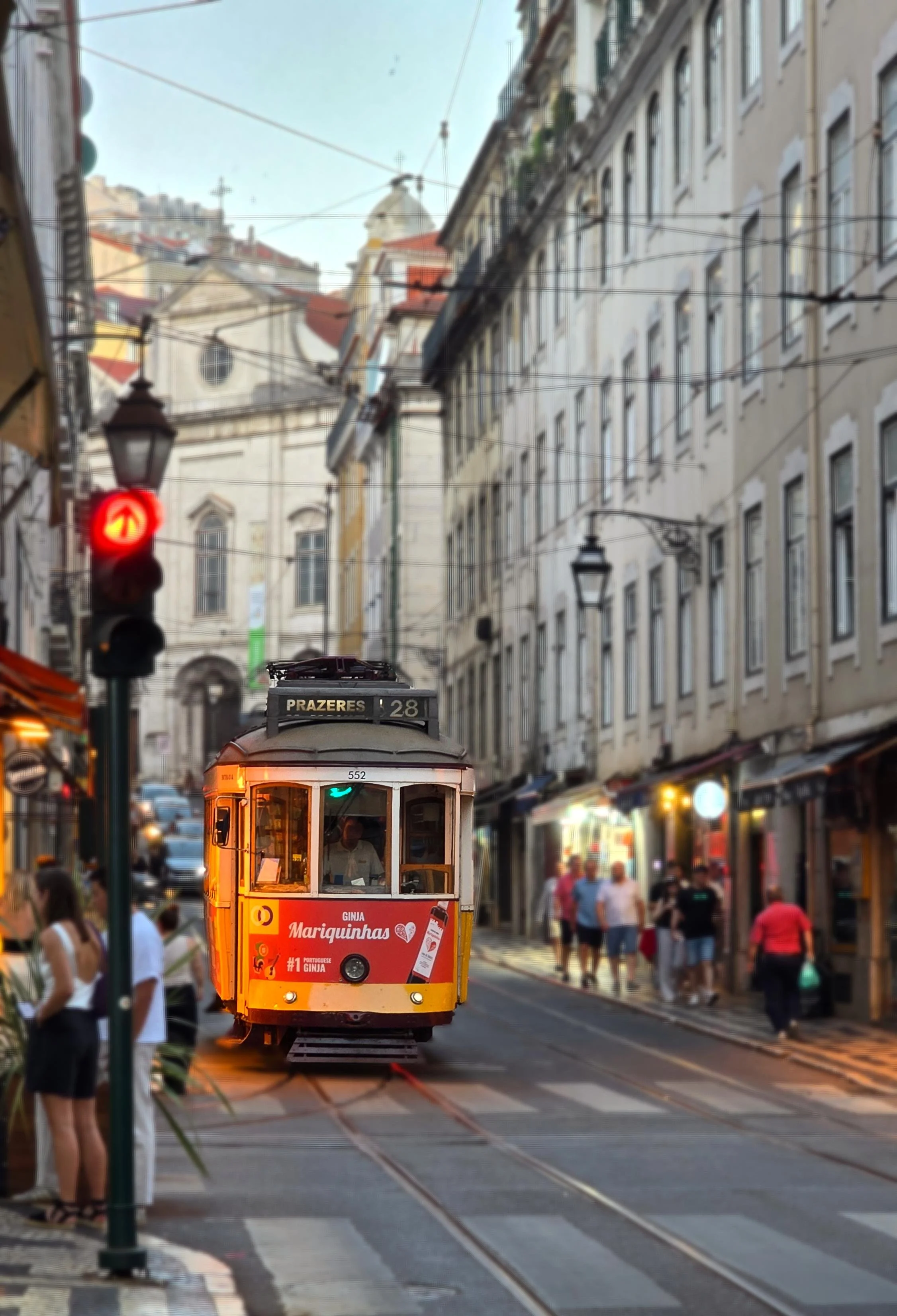 A yellow tram on a city street with pedestrians walking along the sidewalk. There are overhead wires and a traffic light showing red. The street is lined with tall buildings. Trolley in Baixa Neighborhood of Lisbon Portugal