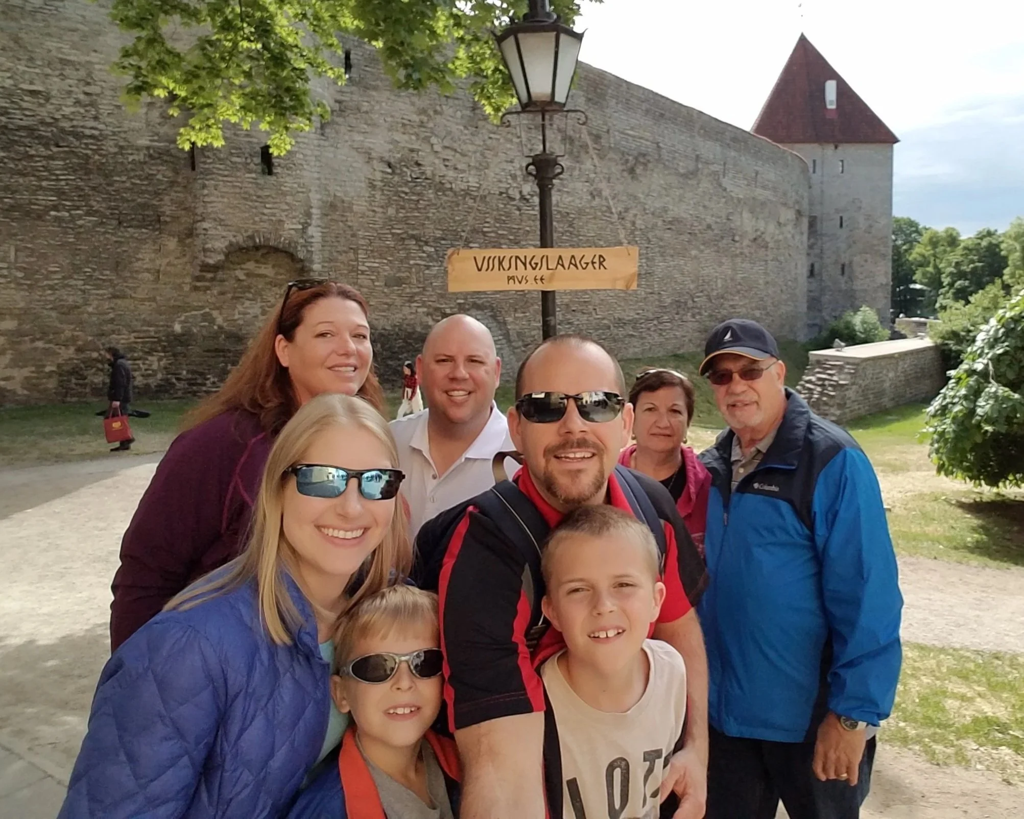 Group of people posing outdoors in front of a stone wall with a sign that reads 'Viskunglaanger'. One woman has sunglasses on her head, and others are wearing sunglasses. There is a woman holding a dog and another person holding a camera. The background includes a historic tower with a red roof and green trees.