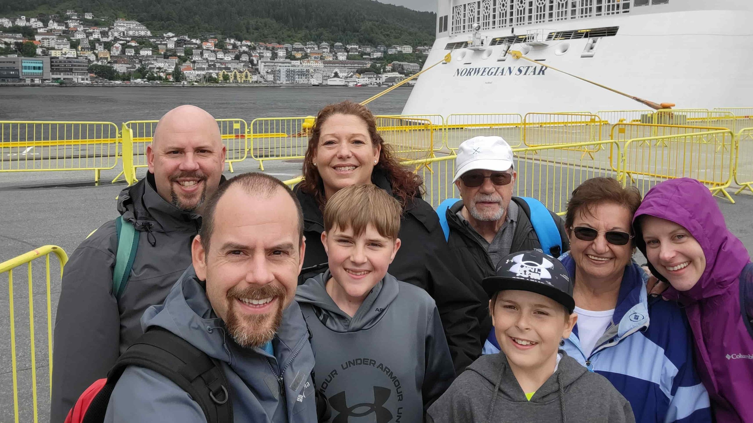 Group of people smiling for a photo on a dock with yellow safety barriers, in front of a white cruise ship named Norwegian Star, with a hillside town in the background.
