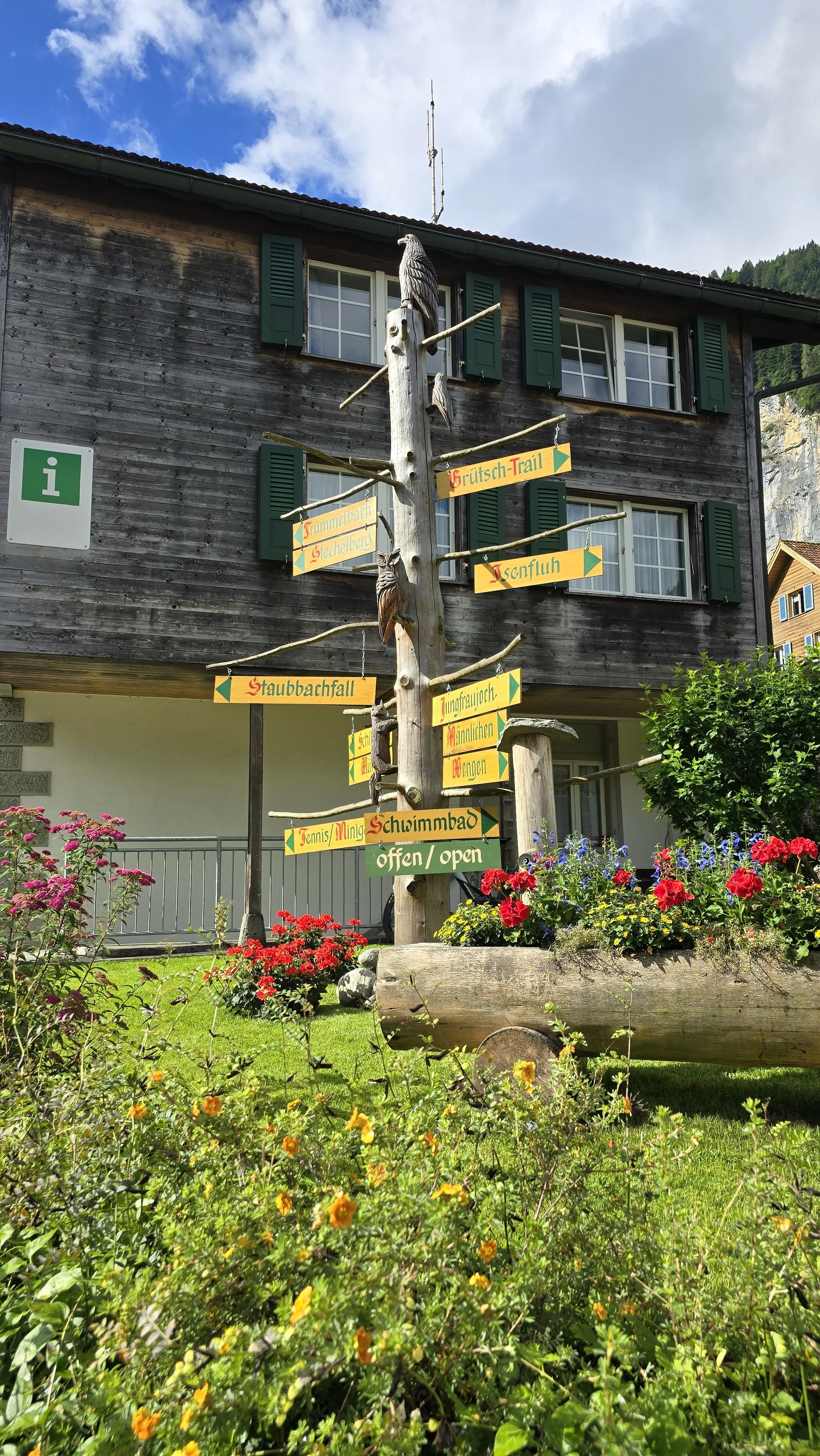 Wooden building with green shutters and a signpost made of a tree branch with multiple colorful signs indicating directions to various trails and activities, surrounded by flowers and greenery.