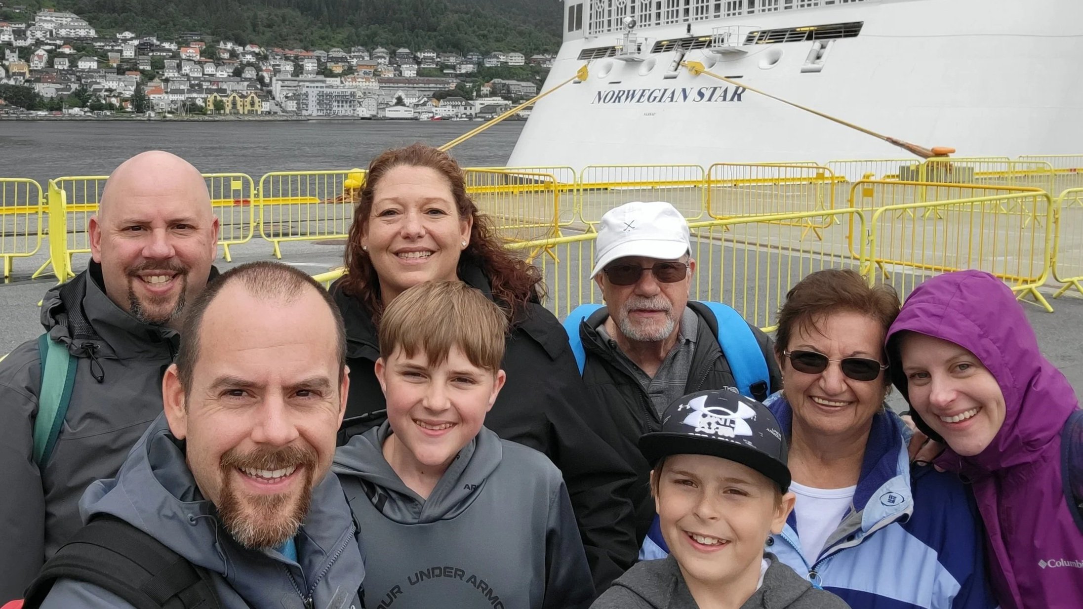 Group of people taking a selfie in front of a large cruise ship named Norwegian Star docked at a waterfront with hillside buildings in the background.