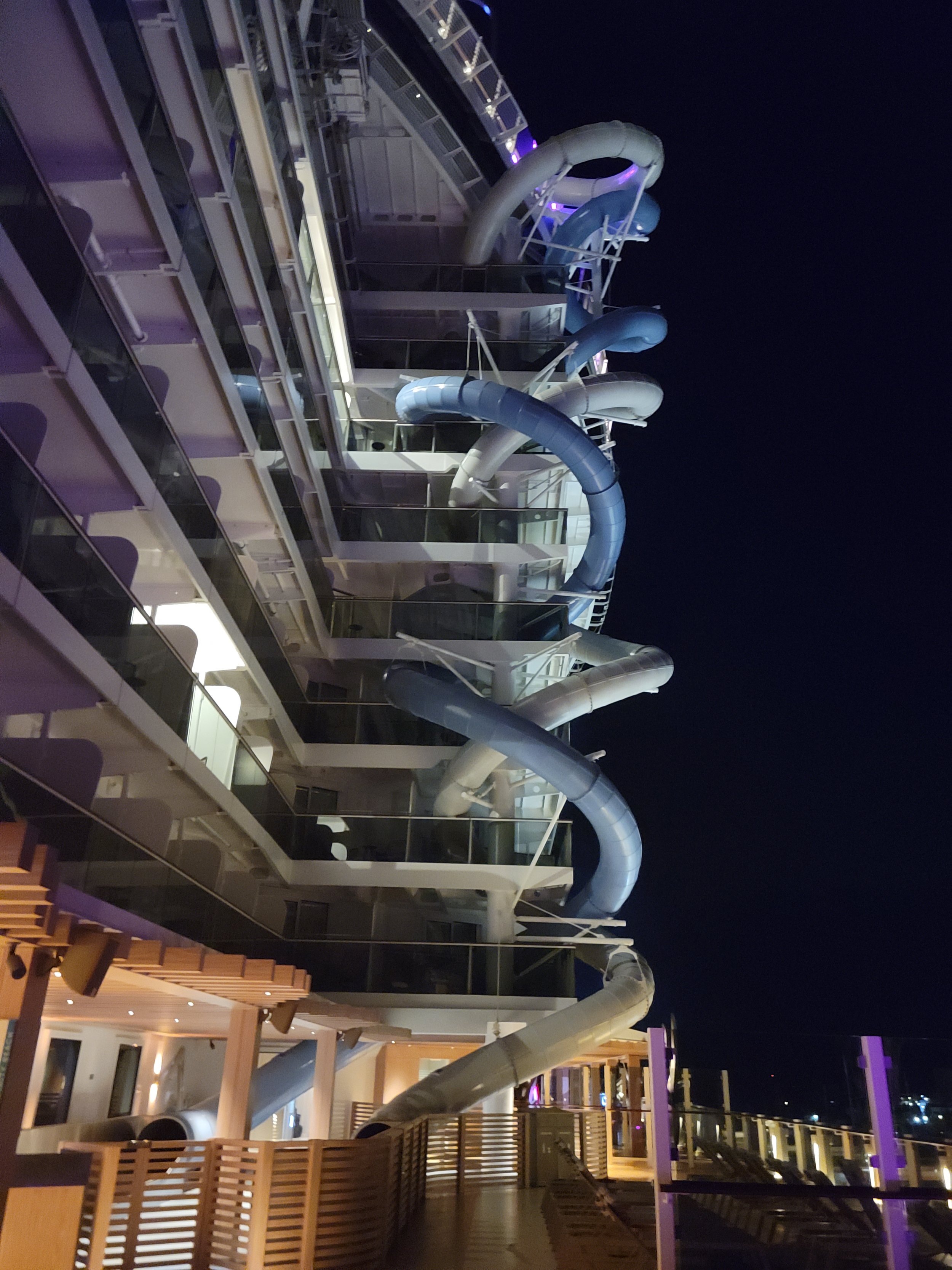 Blue and white enclosed slide tubes spiraling down the side of the Norwegian Prima at night, passing rows of balconies and ending above a wooden-railed outdoor deck.