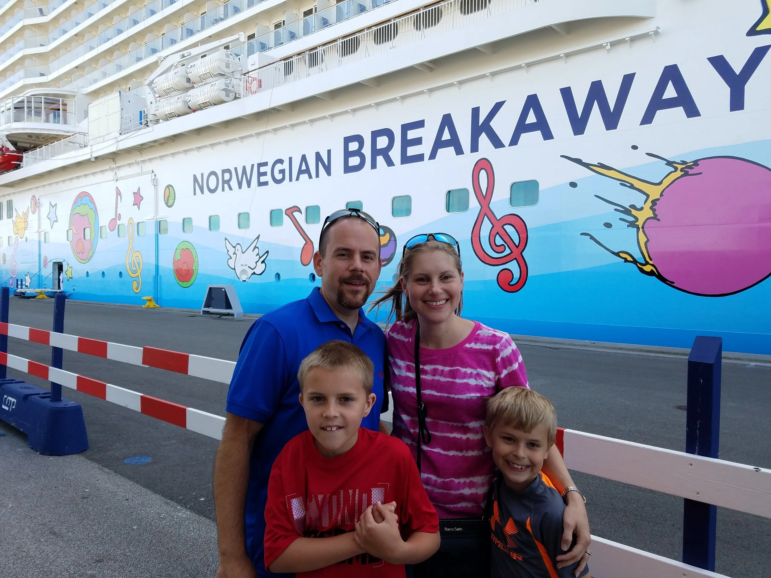 A family of four posing on the pier in front of the Norwegian Breakaway, with the ship's colorful painted hull behind them.