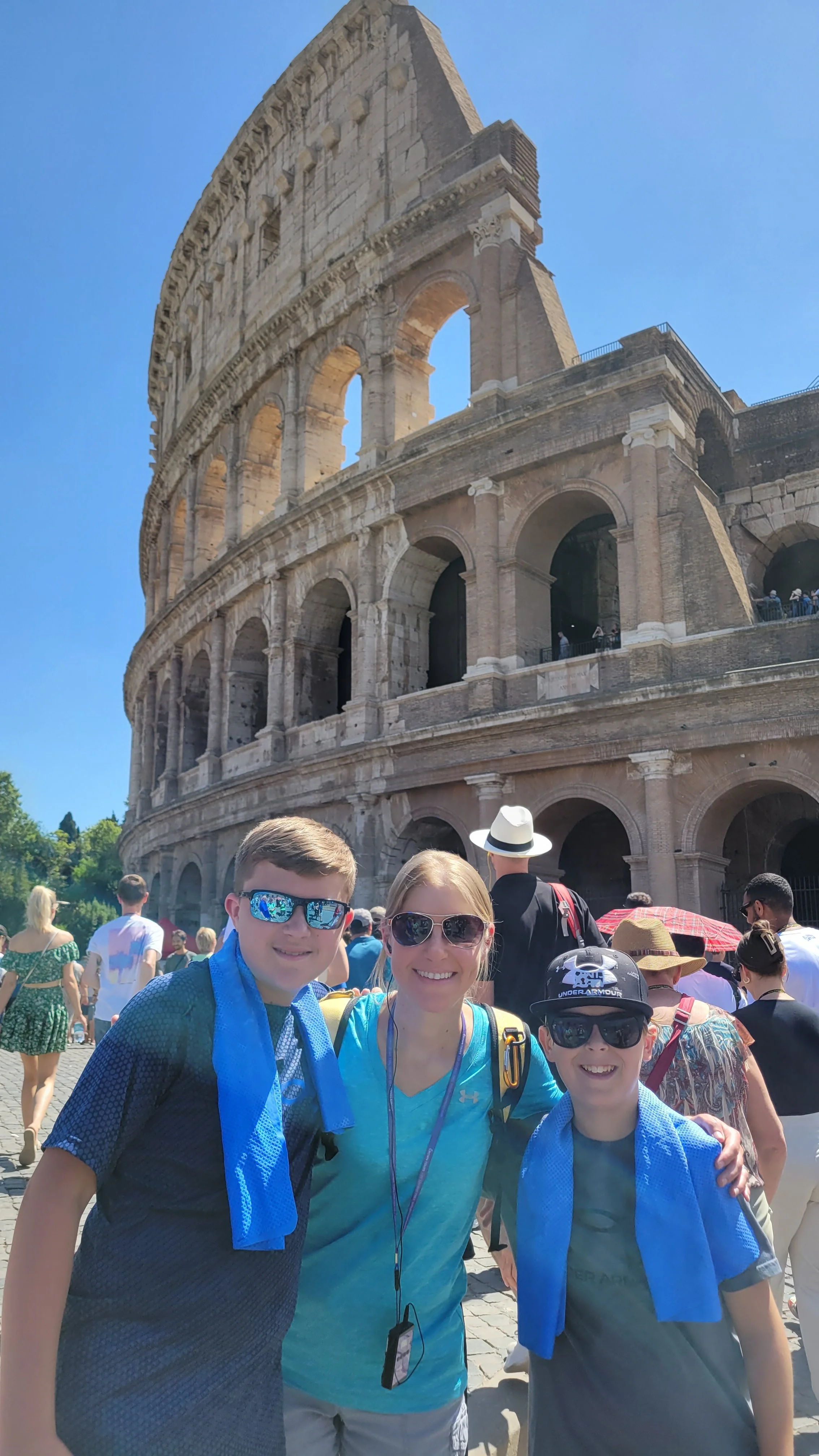 A woman standing between two boys wearing blue cooling towels around their necks, in front of the Colosseum in Rome, Italy, on a sunny day.