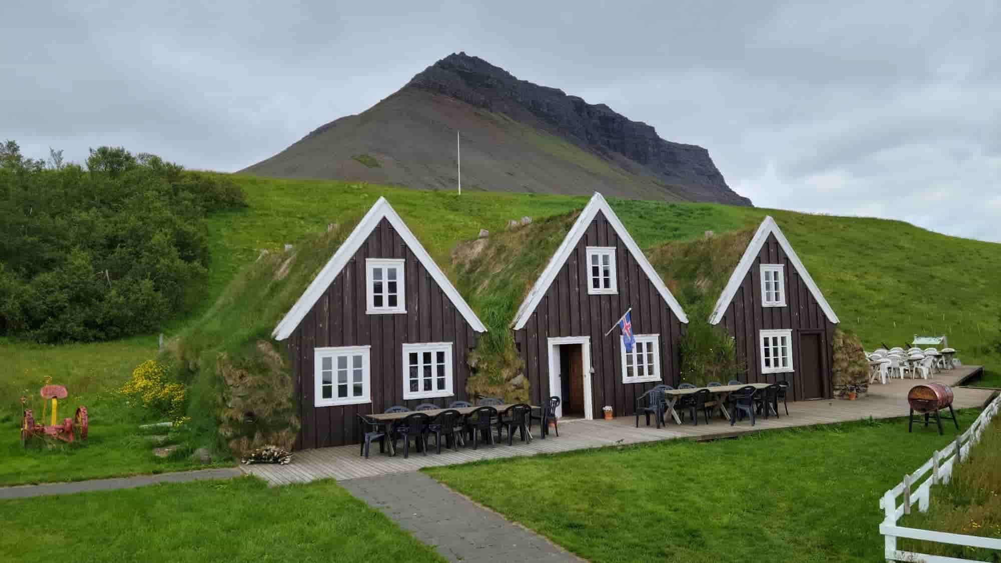 A traditional Icelandic turf house with dark wooden exterior, white window frames, and grass-covered roof, located in a scenic green landscape with a mountain in the background and outdoor seating area.