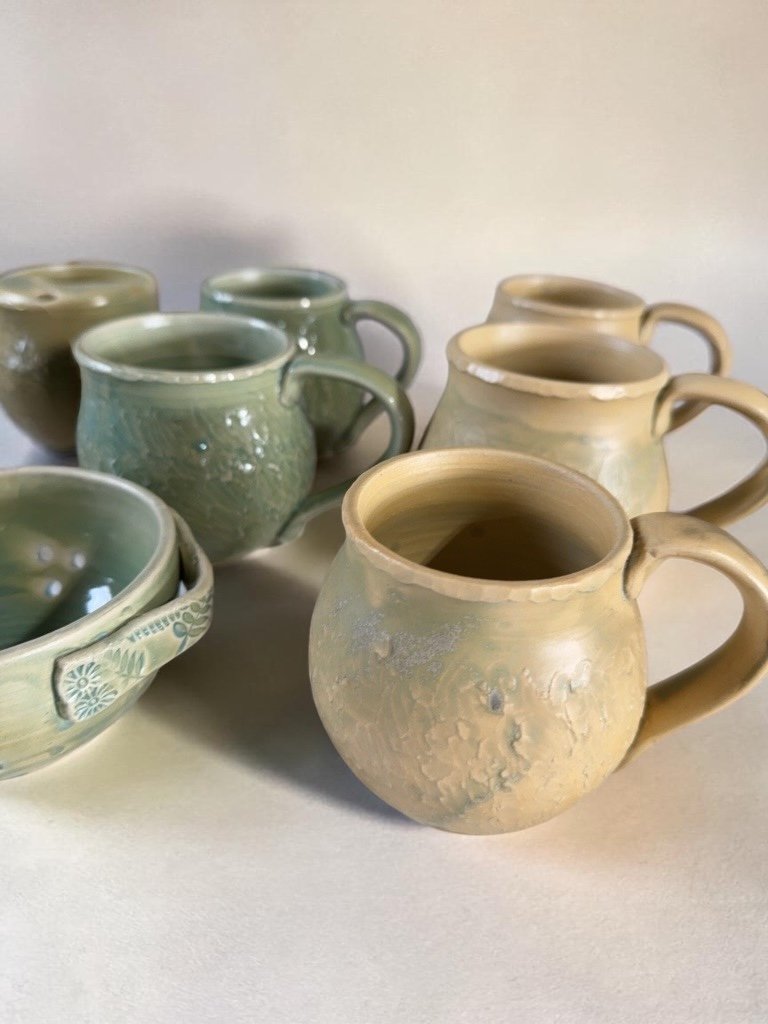 Seven ceramic mugs and a bowl with floral pattern in earthy tones, some mugs are glazed and some have a matte finish, arranged on a plain surface.