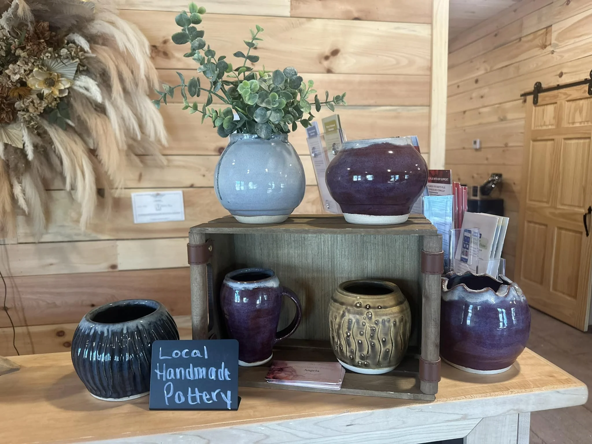 Display of various handmade pottery vases and mugs on a wooden table with a wooden-paneled background, and a handwritten sign reading "Local Handcrafted Handmade Pottery".