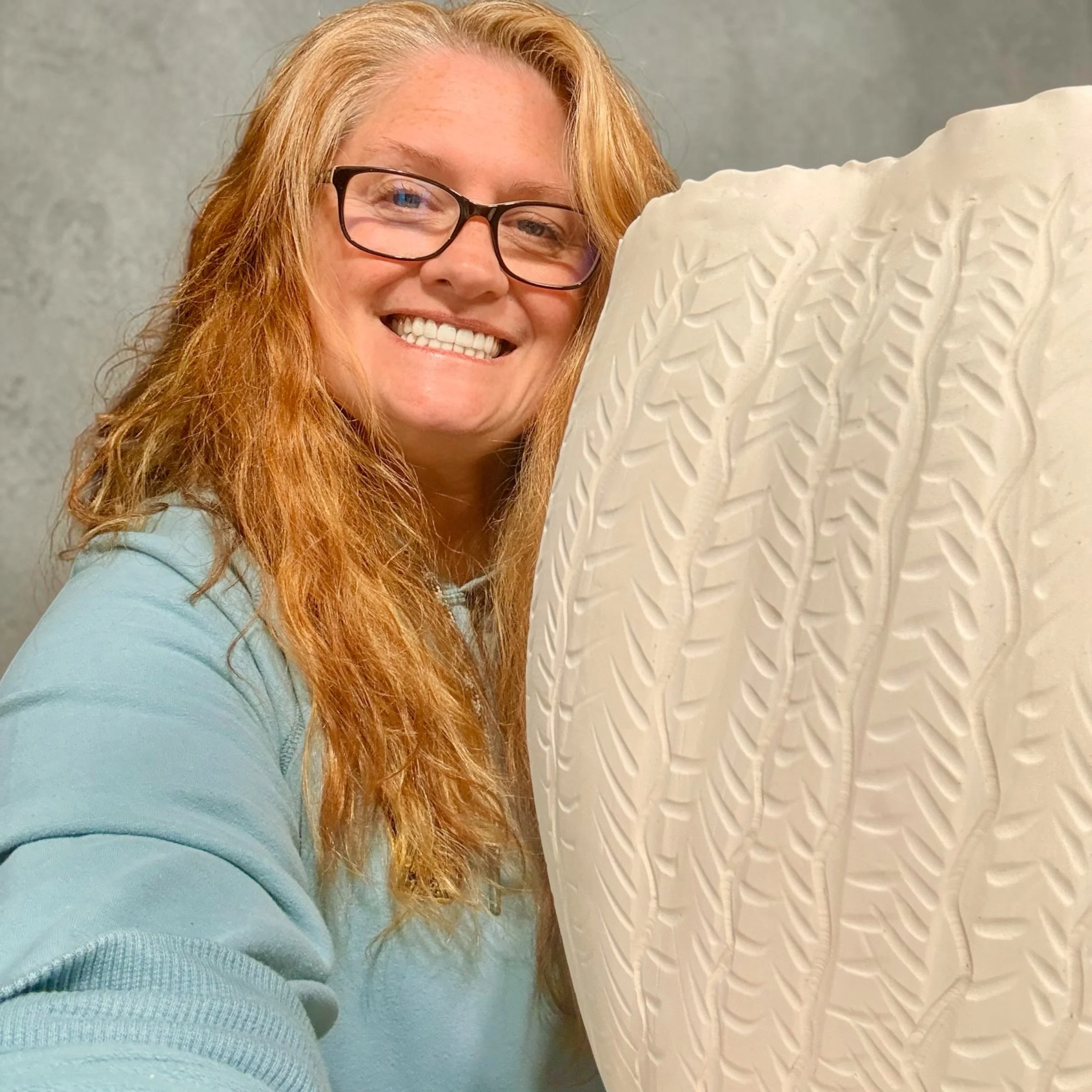 A woman with red hair, glasses, and a bright smile taking a selfie with a large white textured object in front of her, against a plain gray background.