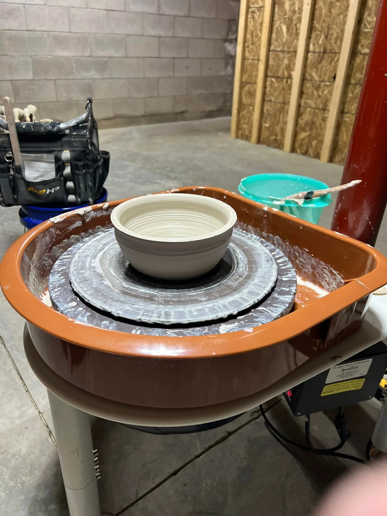 A pottery wheel with a partially shaped ceramic bowl on it, beside a green plastic bowl and a small black tool holder with pottery tools, in a workshop setting with concrete floor and unfinished walls.