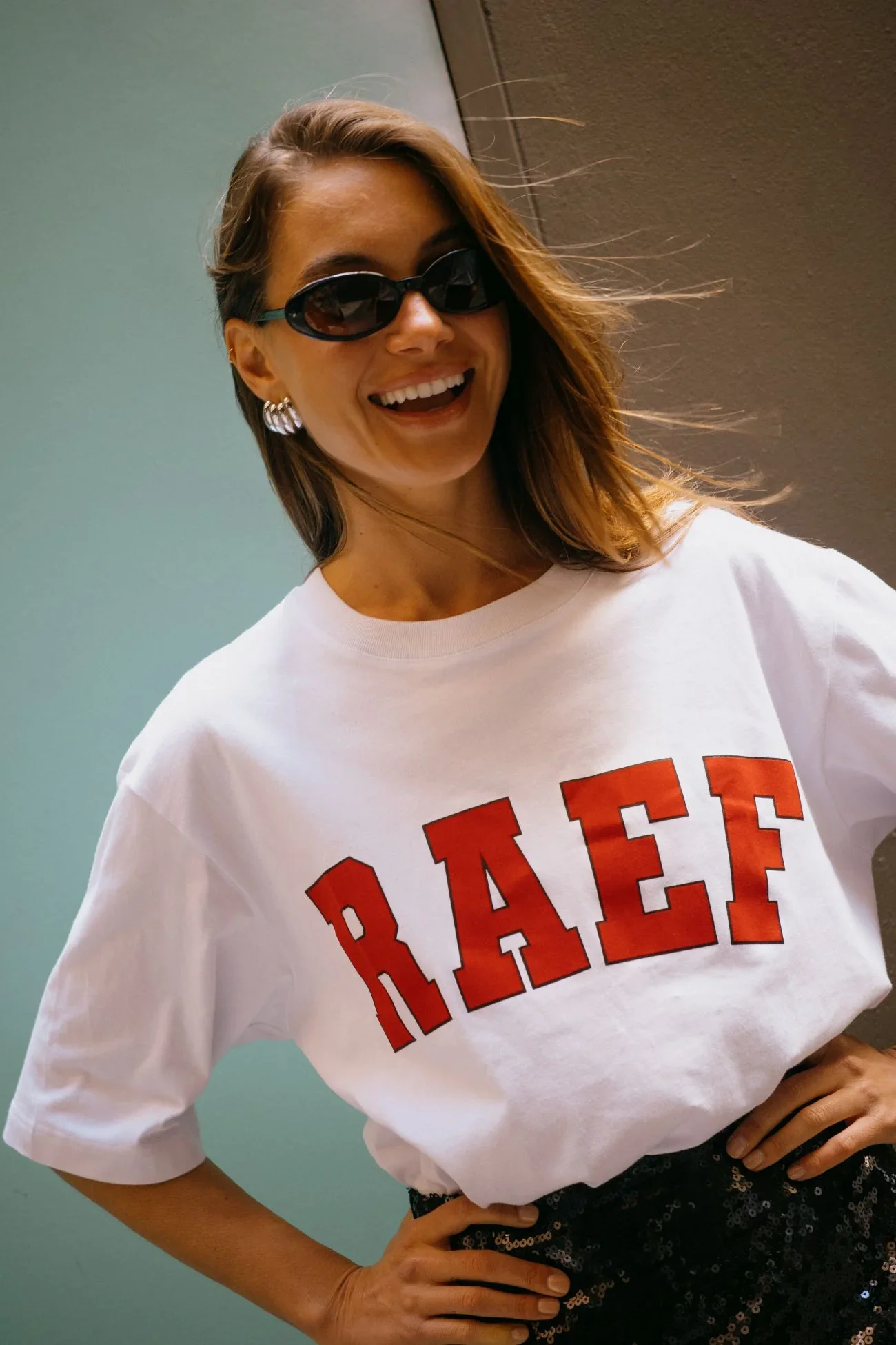 A woman with brown hair and sunglasses smiling, wearing a white t-shirt with red letters and black sequined pants, standing with her hands on her hips against a plain background.
