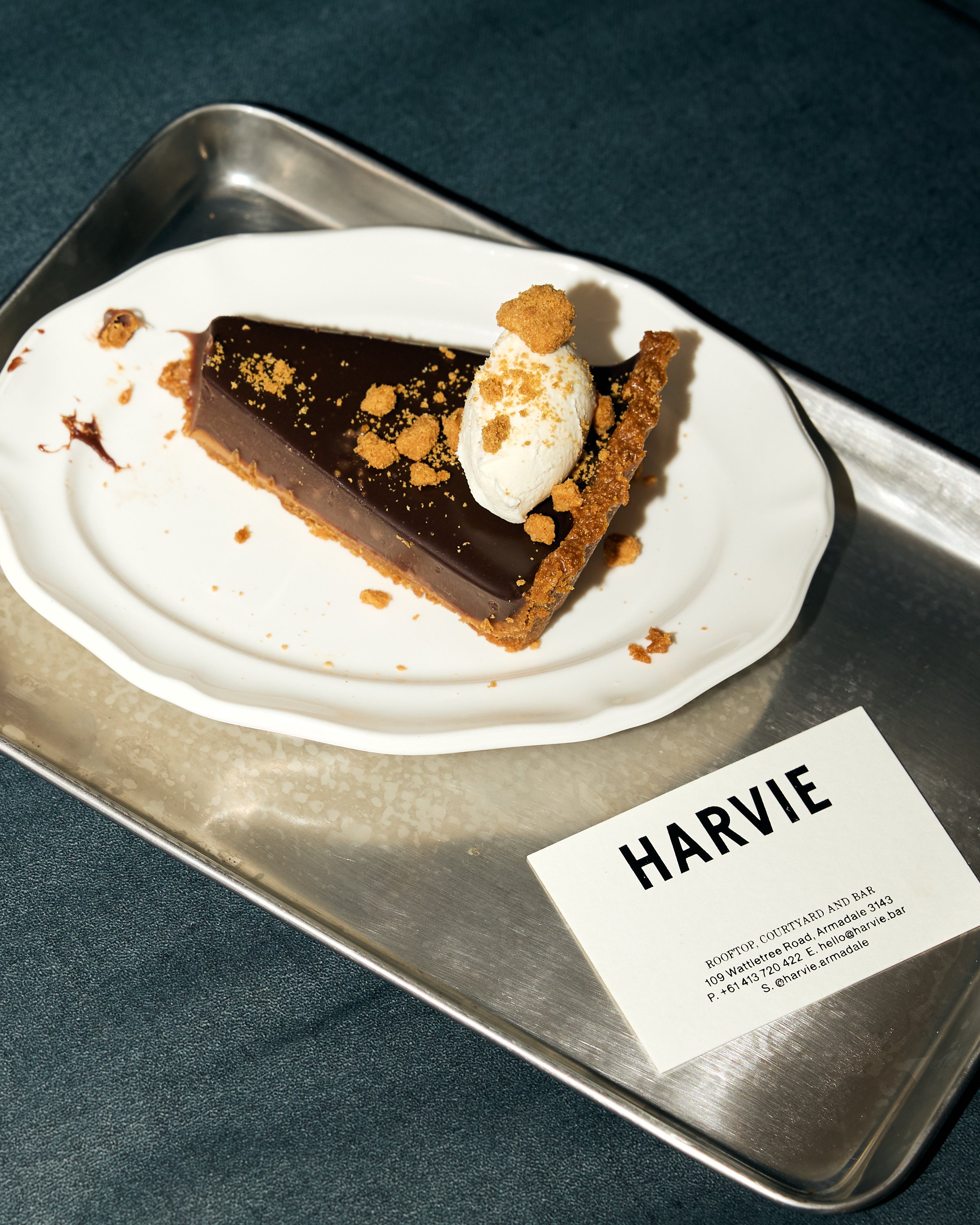 A slice of chocolate dessert on a white plate with a dollop of whipped cream, sprinkled with crumbs, placed on a metal tray, with a name tag reading "HARVIE" underneath on a black surface.