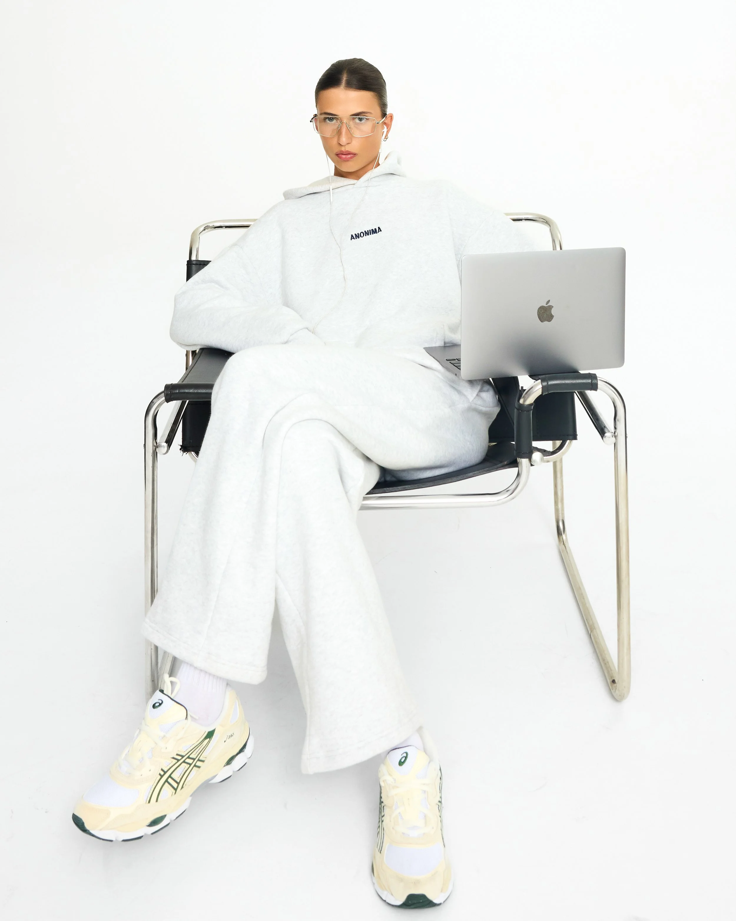 Person sitting in a wheelchair wearing a white hoodie, white pants, and sneakers with a yellow and black design, using earbuds and a laptop on their lap, against a plain white background.