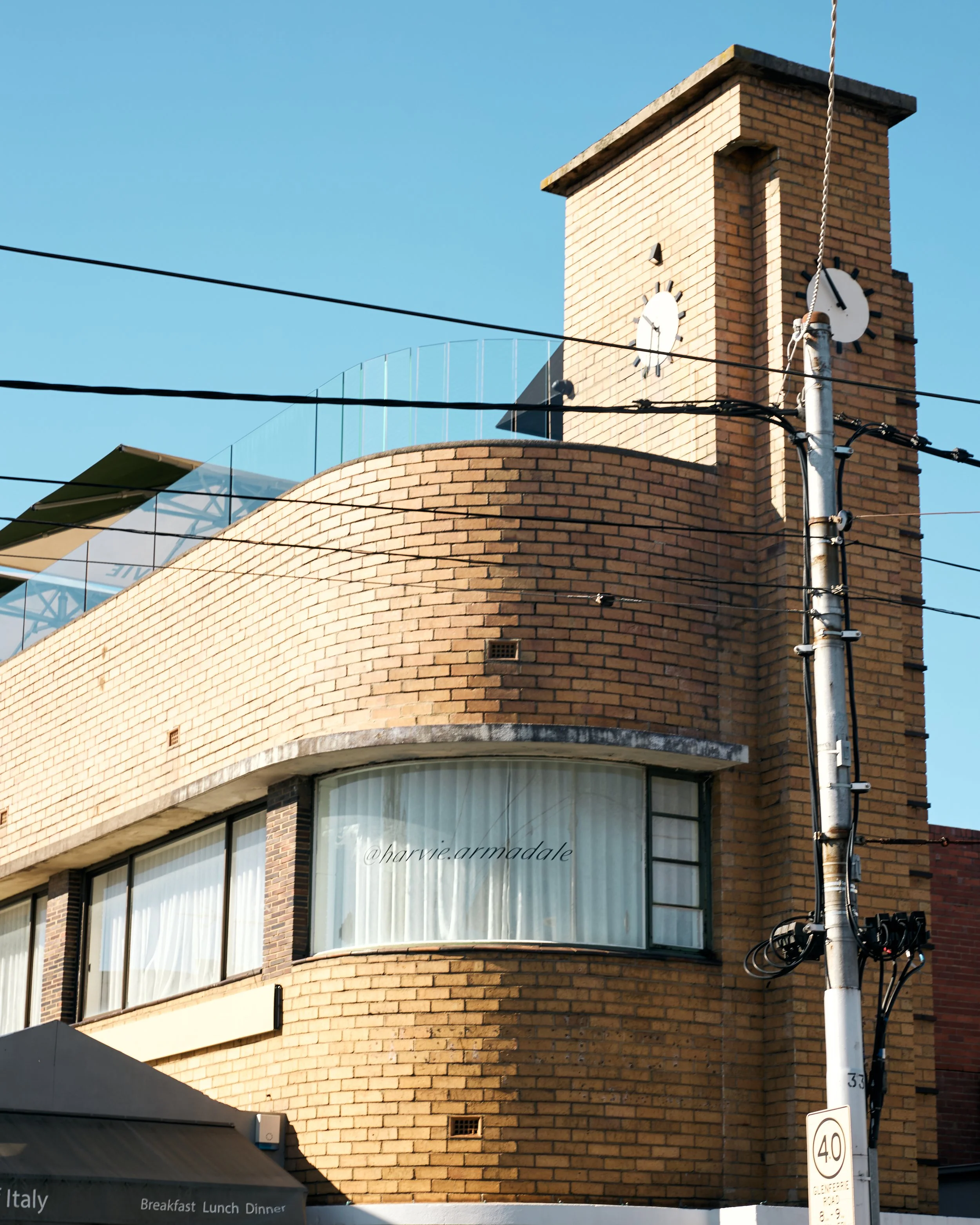Close-up of a beige brick building with a curved facade and large windows. A brick chimney with two clock faces is on the top right, with the time around 7:05. Several black electrical wires and a utility pole are in the foreground. Part of an awning with signage for Italy and breakfast, lunch, dinner is visible at the bottom.