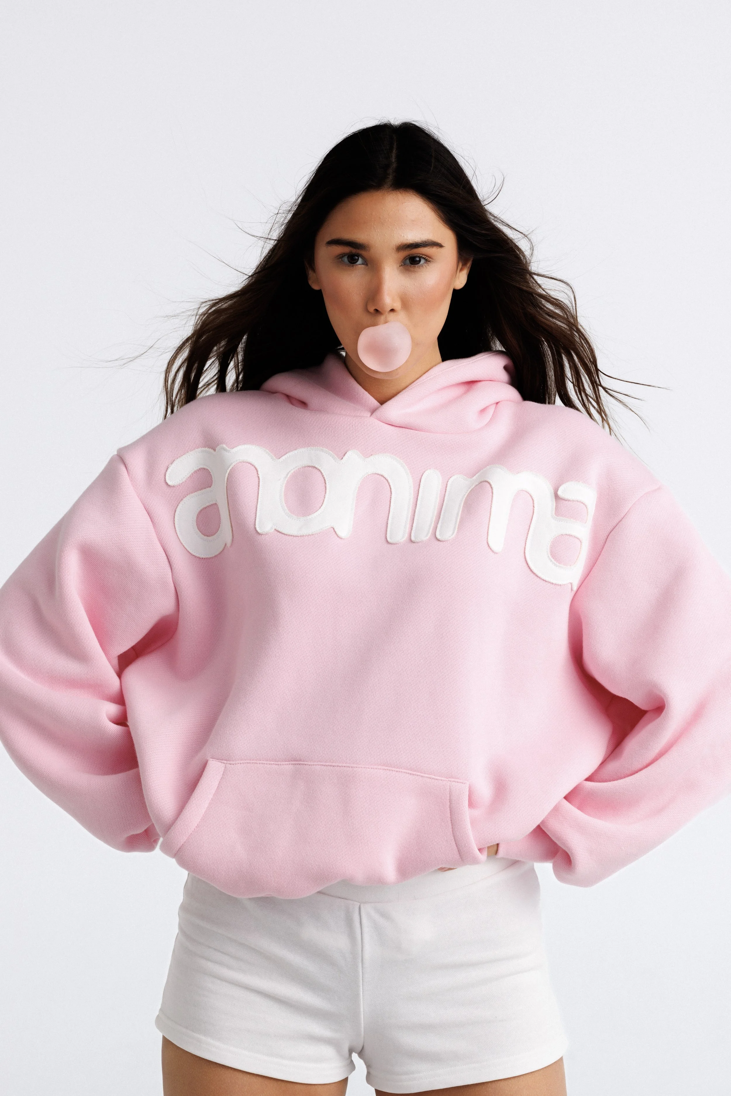 A young woman with long dark hair, wearing a pink hoodie with white letter design and white shorts, is blowing a pink bubble gum bubble.