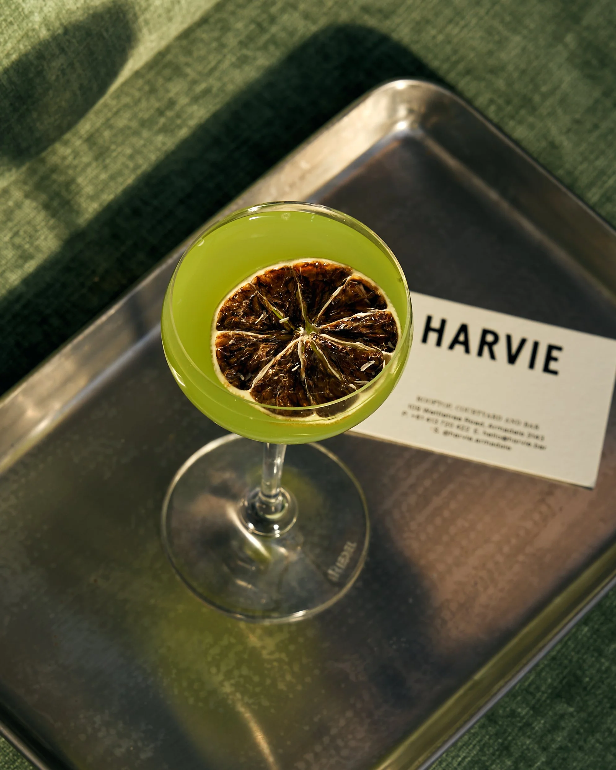 Close-up of a cocktail glass with a green drink and a dried lemon slice garnish, placed on top of a silver tray near a book and a table with a green fabric surface.