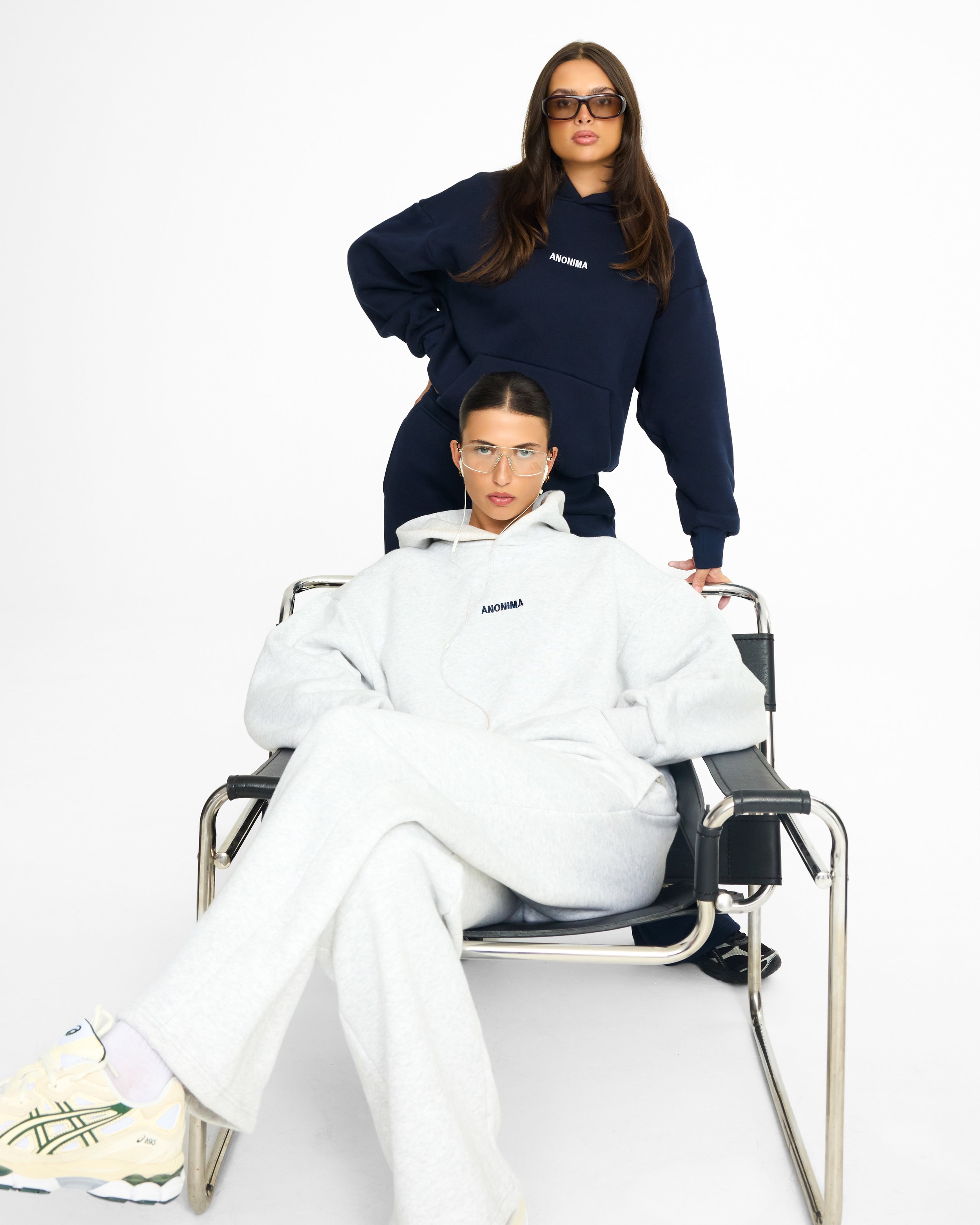 Two young women with glasses, one wearing a navy blue hoodie and the other in a white hoodie, are posing against a plain white background. One woman is standing with her hand on her hip, and the other is sitting in a wheelchair, looking directly at the camera.