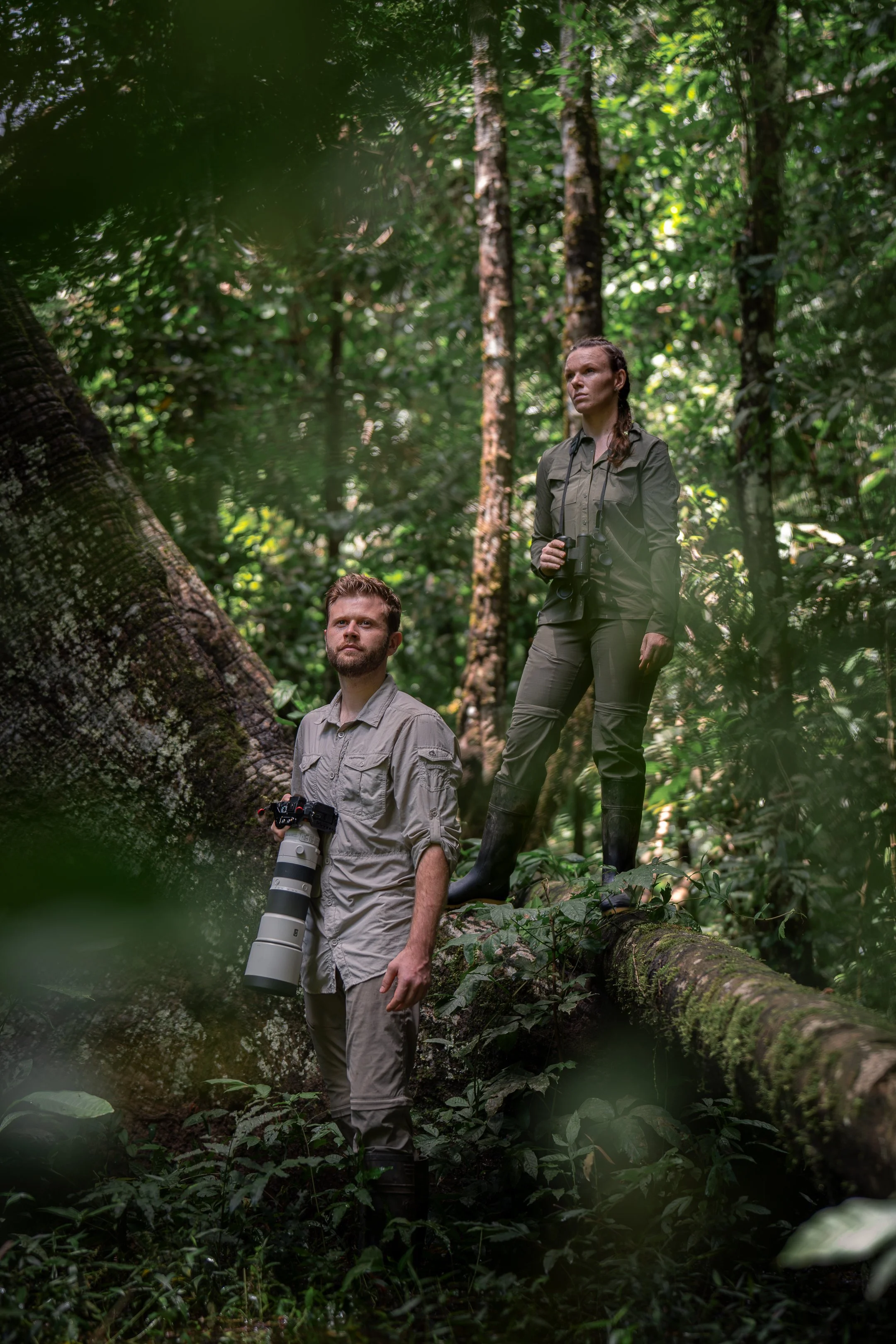 Two people, Luke Pembroke and Nadine Holmes, in a dense jungle of the amazon lowland rainforest; one man sitting on a fallen tree holding a camera with a large lens (Sony 200-600m), and one woman standing with binoculars, looking into the distance.