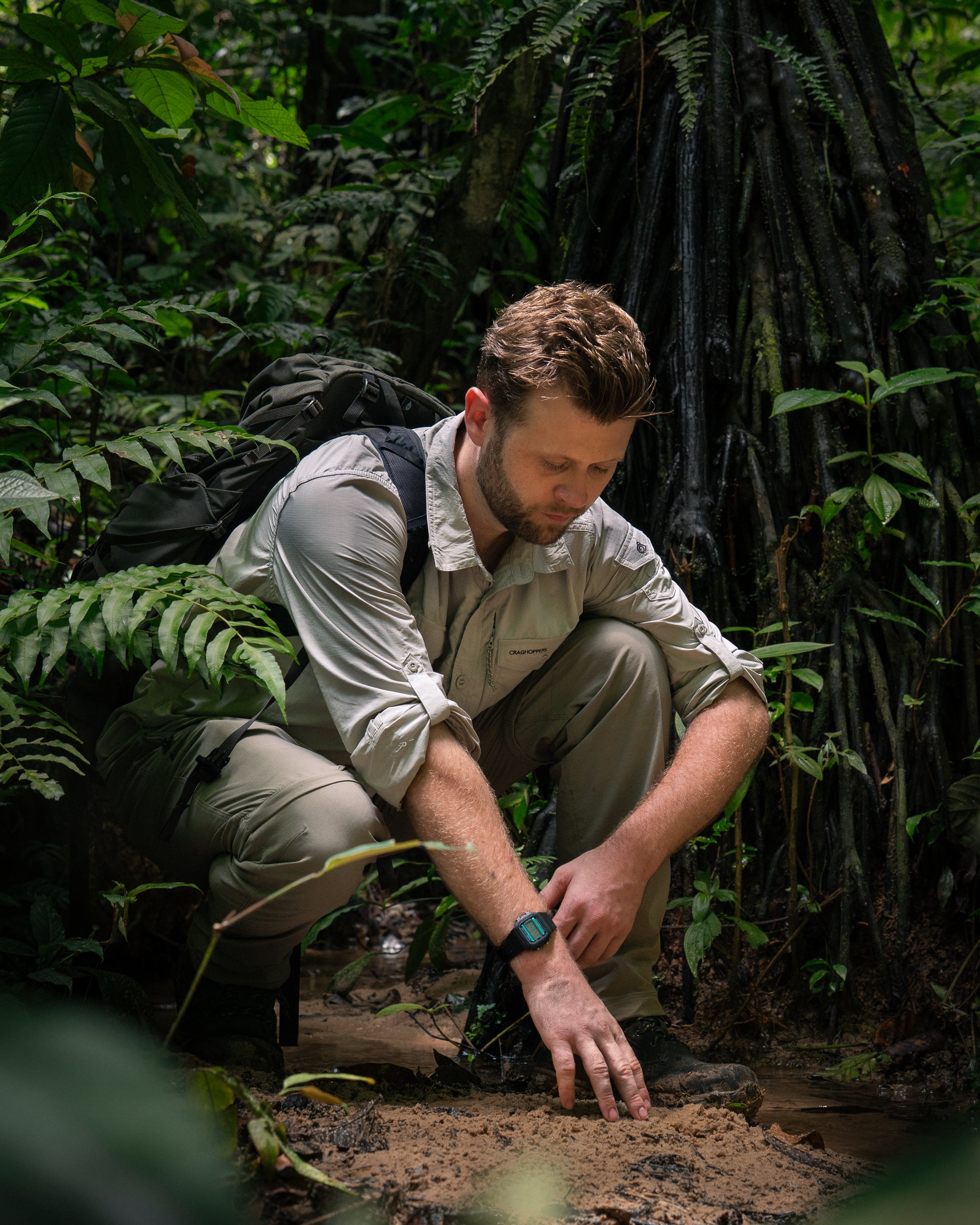 Luke Pembroke, a man with a beard and wearing a beige shirt from Craghoppers and pointing at the water's surface with his right hand, while looking down at the water in the lowland amazon rainforest in Peru 