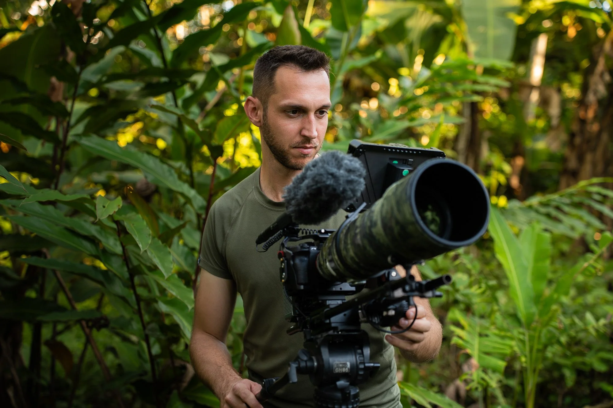 Marc Ferrum, a man with short dark hair and a beard wearing a green t-shirt operating a professional camera with a large Sony 200-600mm telephoto lens in a lush green outdoor environment of the Peruvian lowland amazon rainforest.