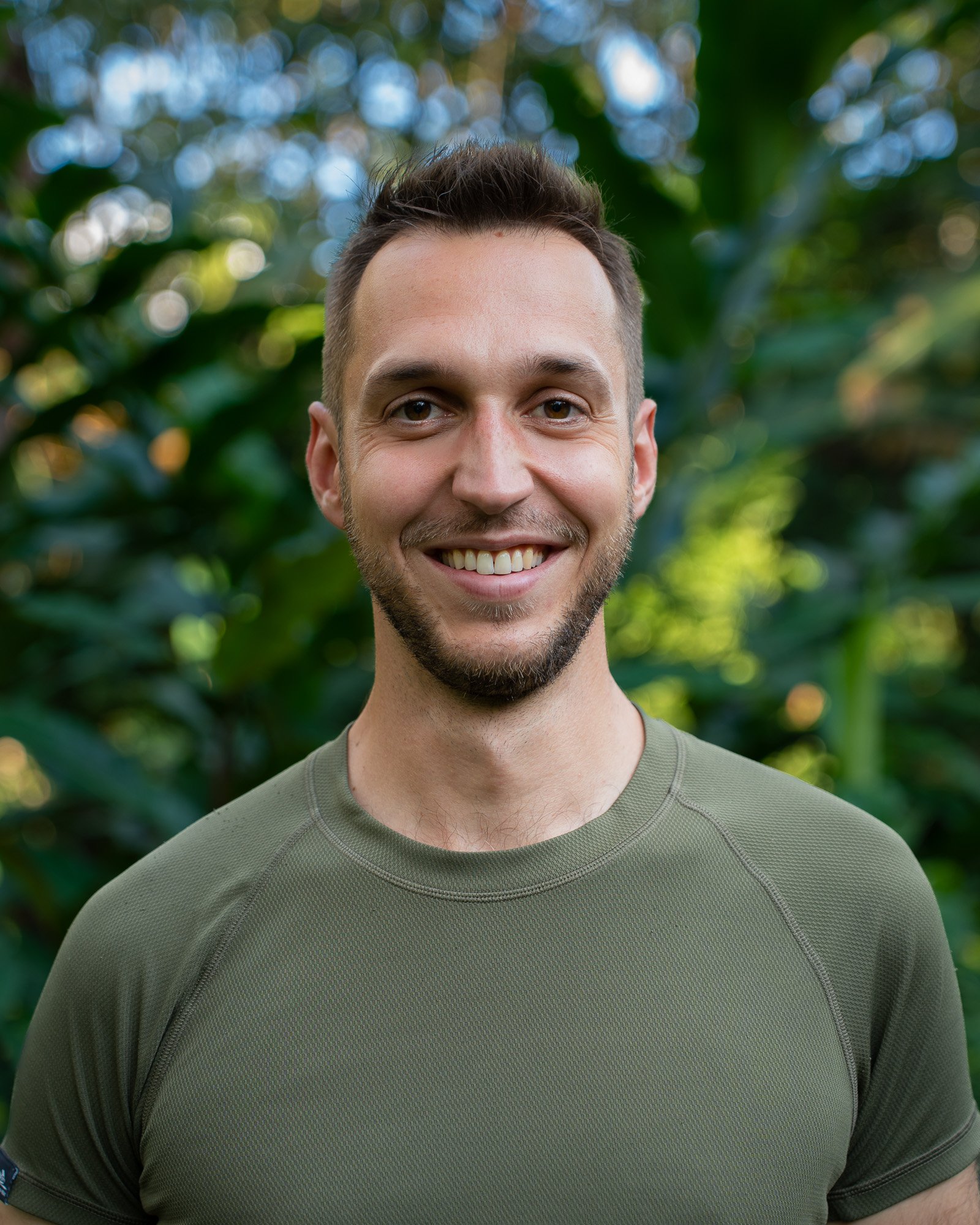 Close-up of Marc David Ferrum, a smiling young man, with short brown hair, a beard, and brown eyes, wearing a green athletic shirt, standing outdoors with green foliage blurred in the background in the peruvian Lowland Amazon Rainforest