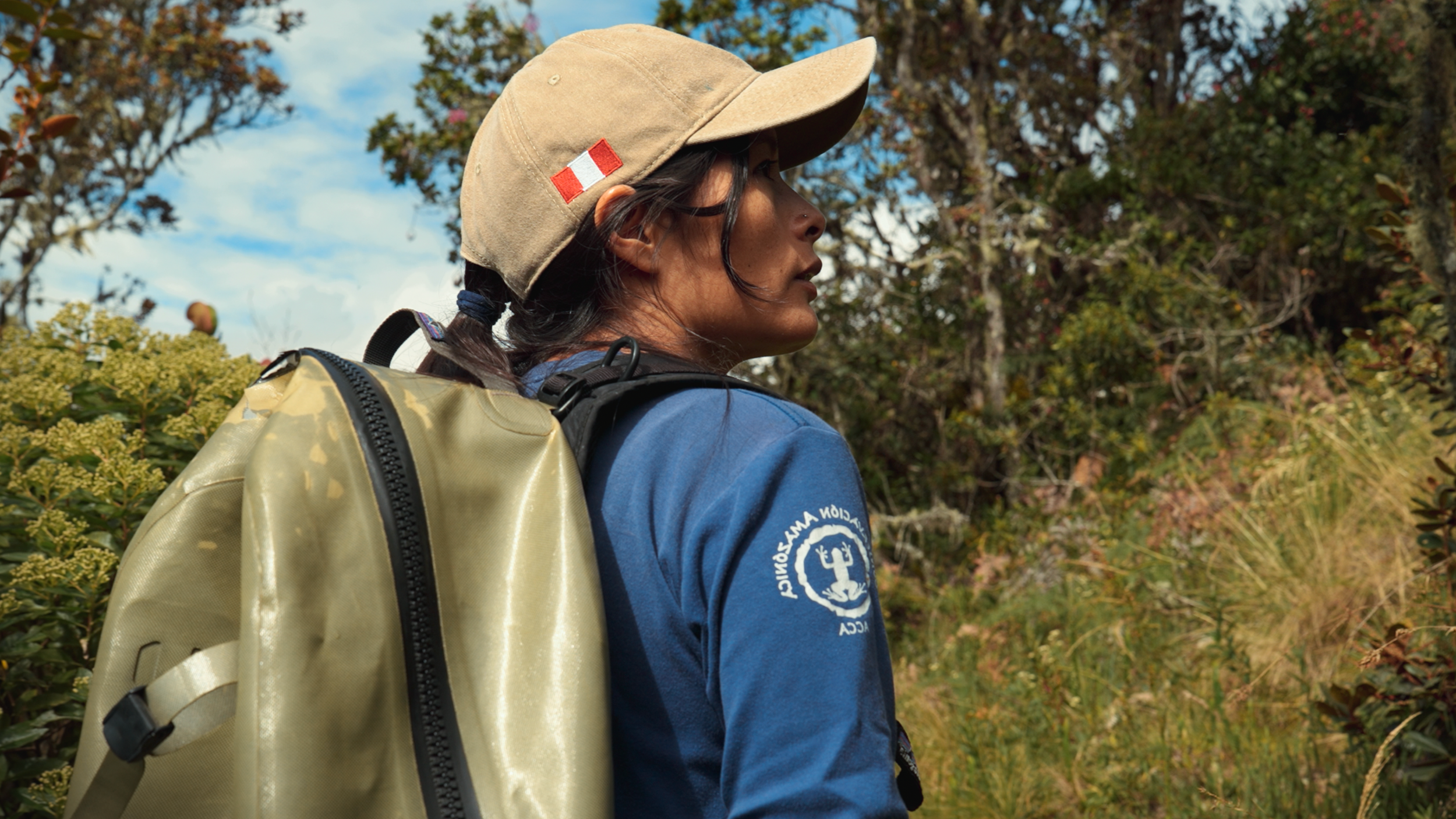 Ruthmery Pillco Huarcaya, an Andean woman, wearing a tan hat with a red and white patch, a blue hoodie with a logo, and a tan backpack, standing in the Peruvian cloud forest at Wayqecha surrounded by green bushes and trees under a partly cloudy sky.
