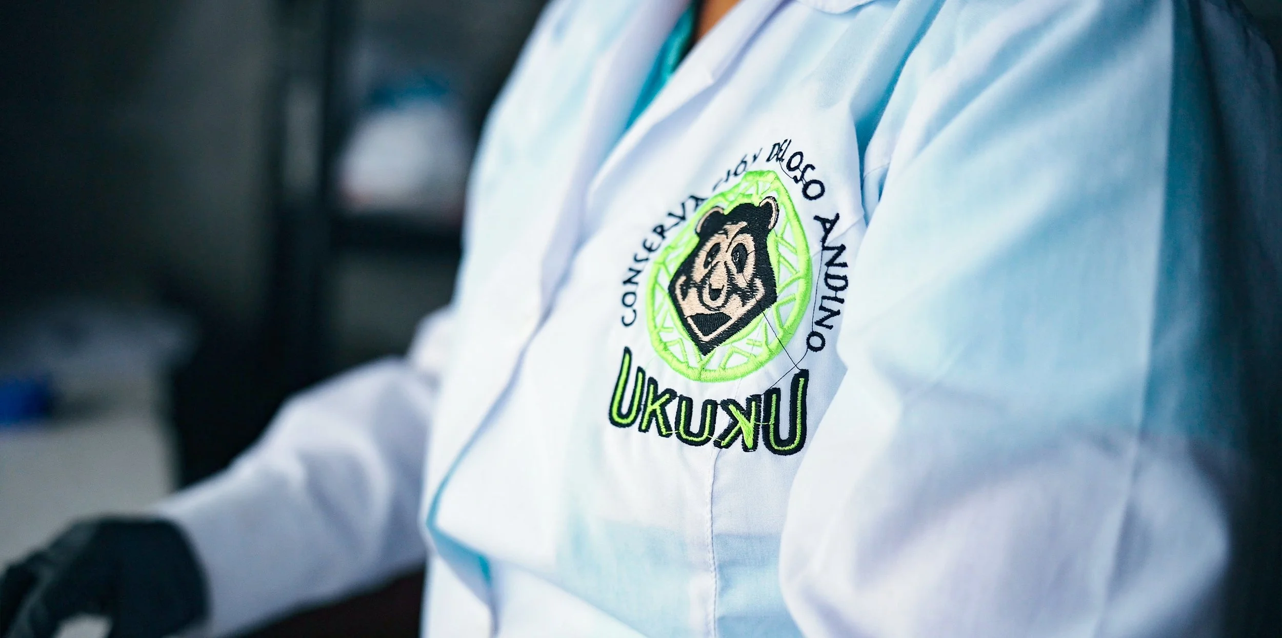 Close-up of a person wearing a white jacket with a logo featuring a bear's face and the text 'Conferencia Mundial Anual Ukuku' embroidered on it, with the person's arm and part of their torso visible at Wayqecha Biological Station