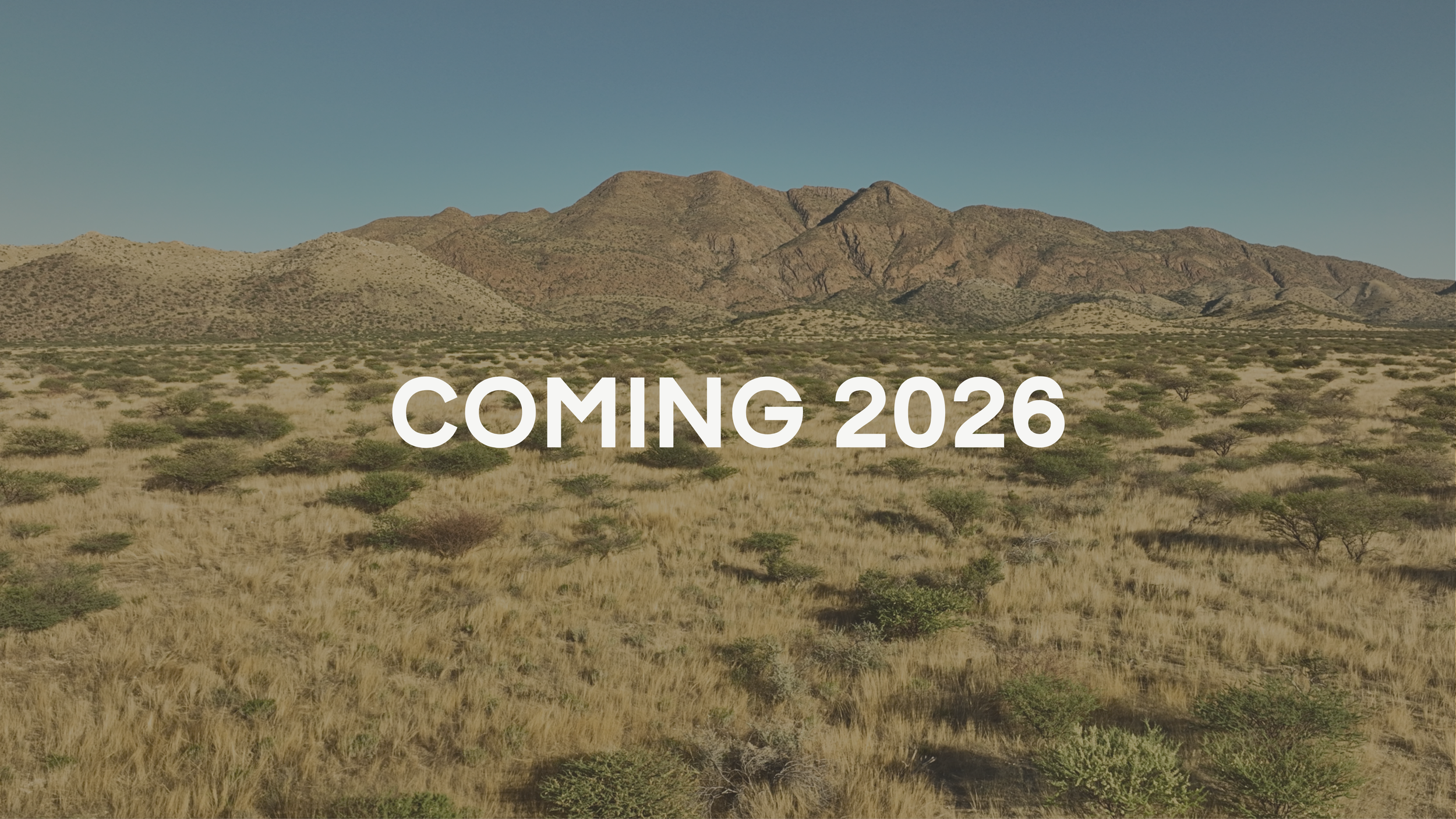 A desert landscape with mountains under a clear sky, with the text 'COMING 2026' overlayed. Photographed at Etusis Lodge in the Erongo Region of Namibia.