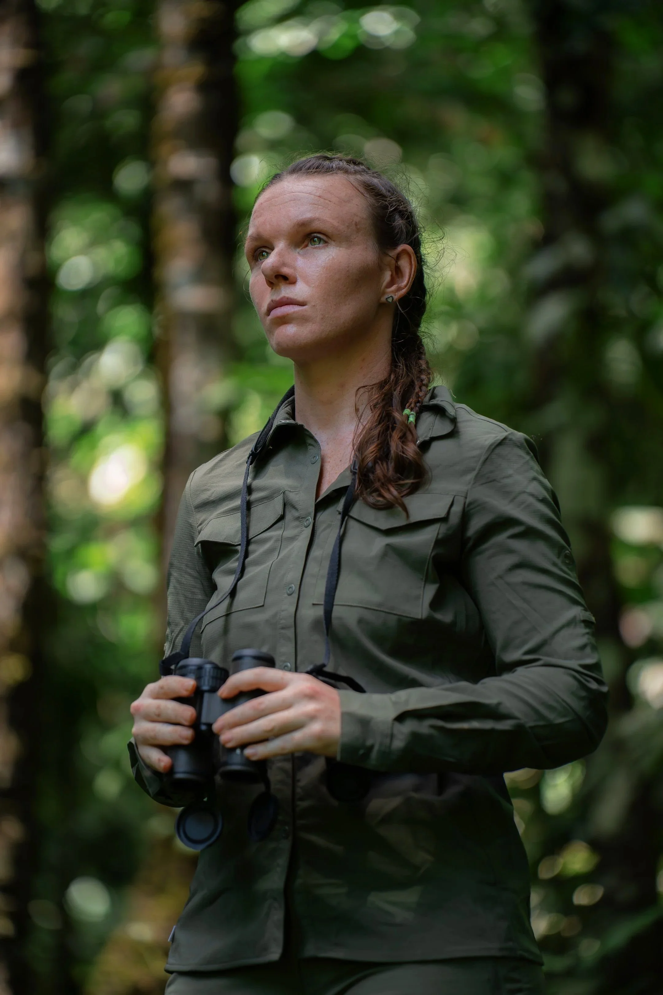 Nadine Holmes, a woman with braided hair and light skin wearing a hiking jacket from Craghoppers holding binoculars, standing in a forest with sunlight filtering through trees in the lowland amazon rainforest of Peru with Fauna Forever