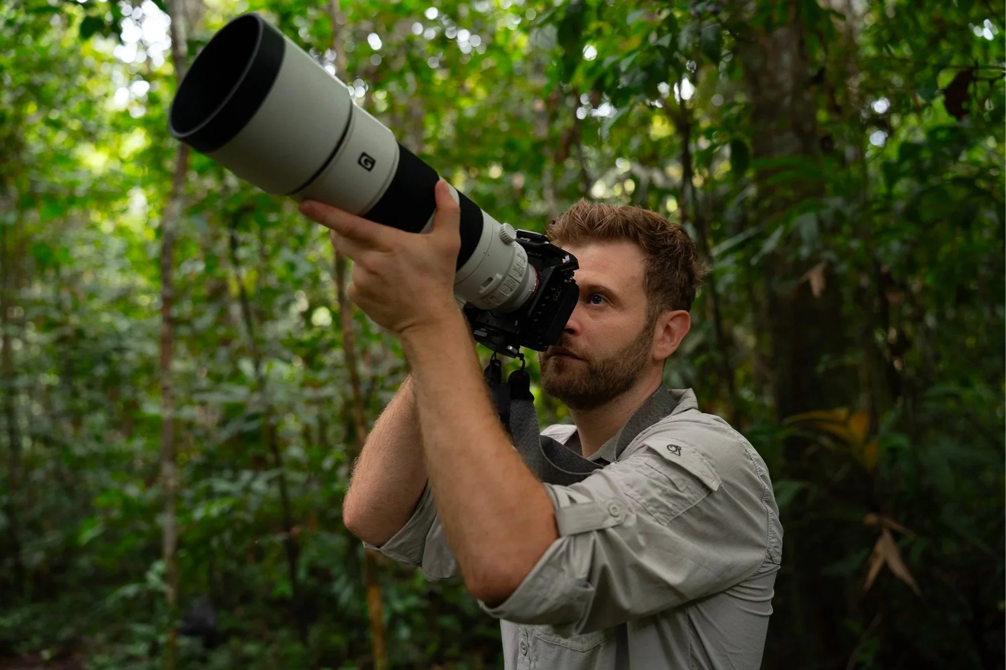 Luke Pembroke, a man with a beard and short hair taking a photograph with a large Sony 200-600mm telephoto lens in a dense green forest of the Peruvian Amazon Rainforest.