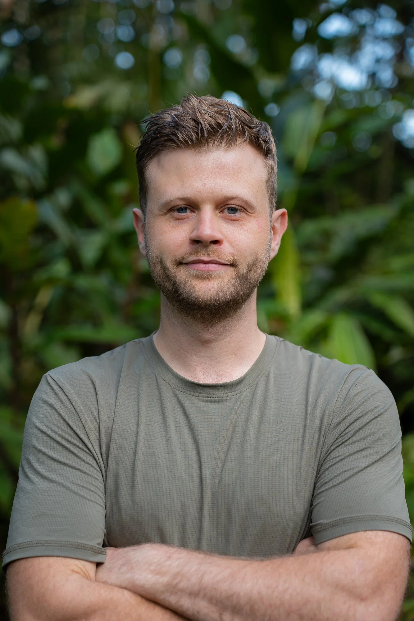 Luke Pembroke, a man with short brown hair and a beard, smiling softly with crossed arms, in front of a blurred green foliage background of the Peruvian lowland Amazon Rainforest.