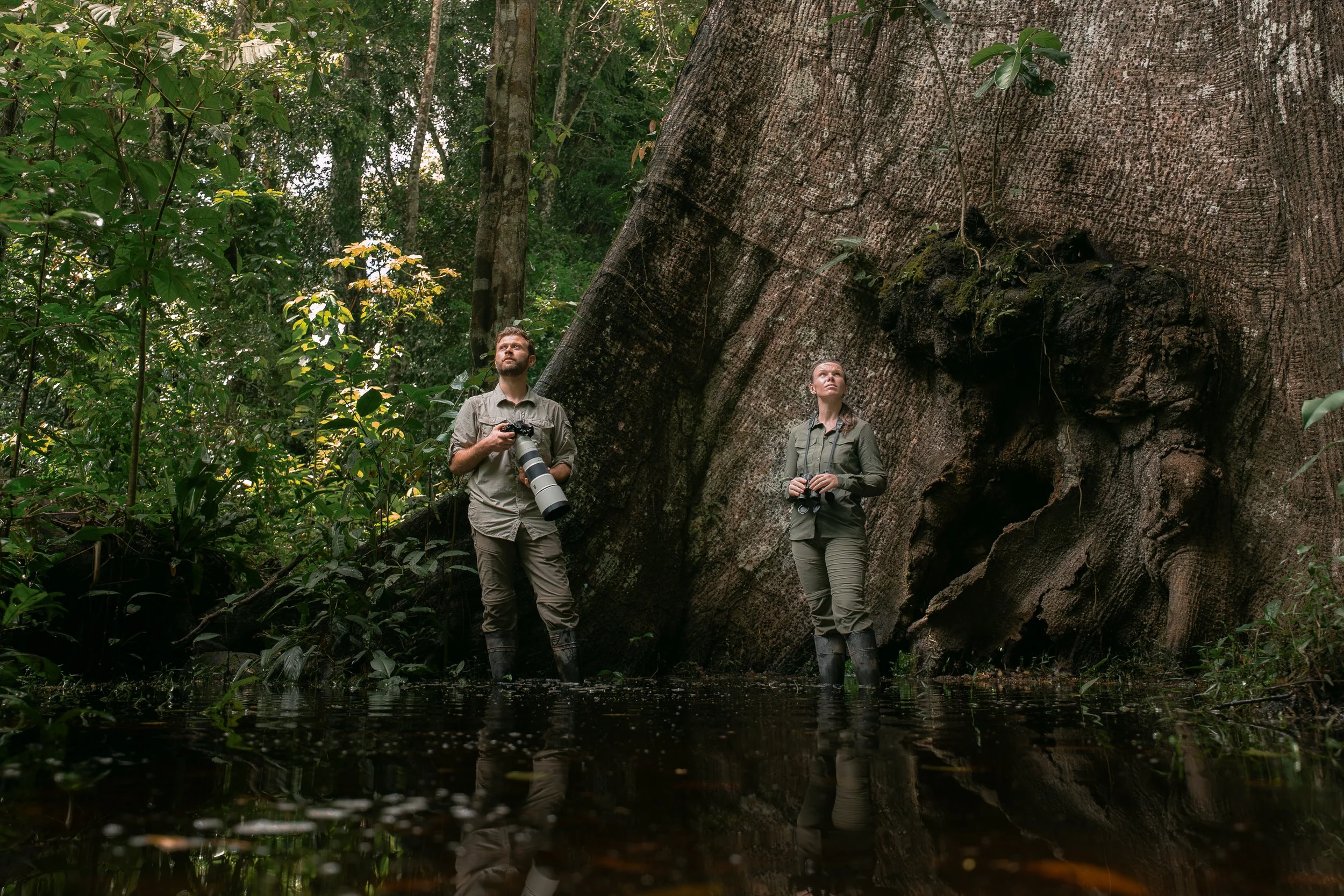 Two people, Nadine Holmes and Luke Pembroke, a man and a woman, standing in a lush, green rainforest next to a large tree. They are holding binoculars and a camera with a large telephoto lens, looking up at the canopy of the lowland amazon rainforest