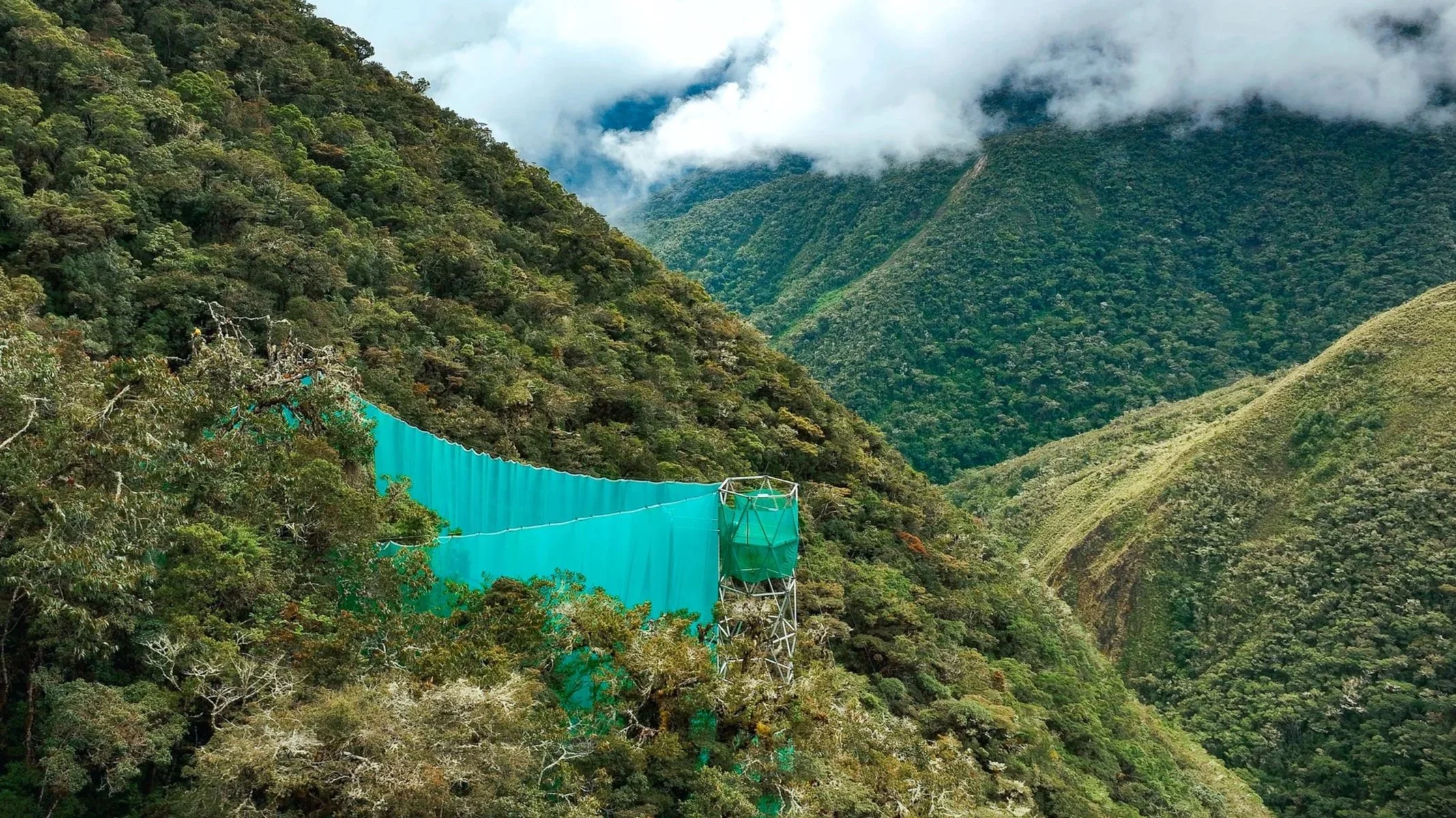 A scenic view of lush, green mountains with a cloudy sky overhead and a turquoise-colored bridge or dam structure in the foreground at Wayqecha Biological Station in the high Andean cloud forest of Peru. 