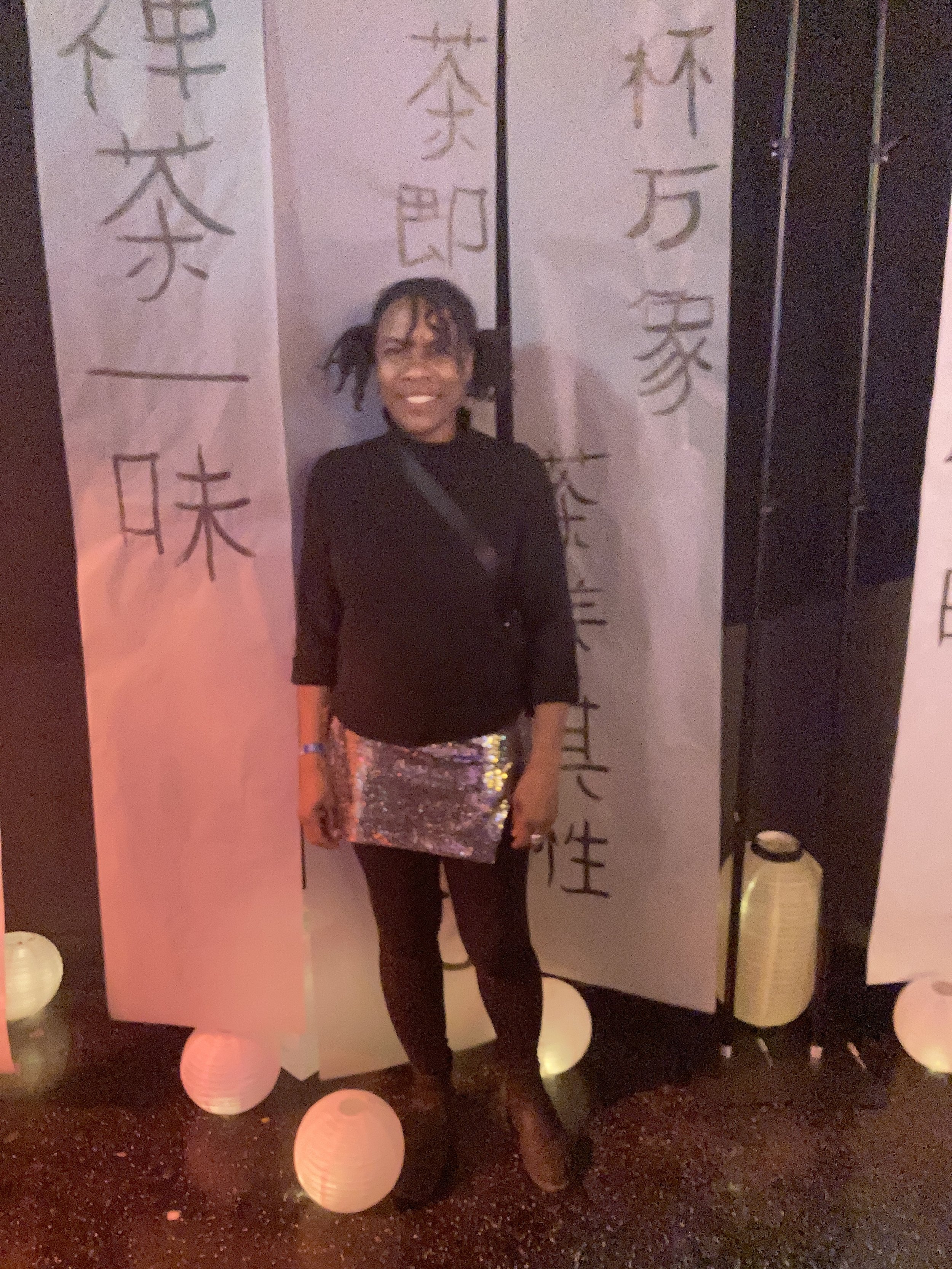 A woman smiling and standing in front of a Japanese calligraphy backdrop at an indoor event. She is wearing dark clothing and a shiny, reflective skirt.