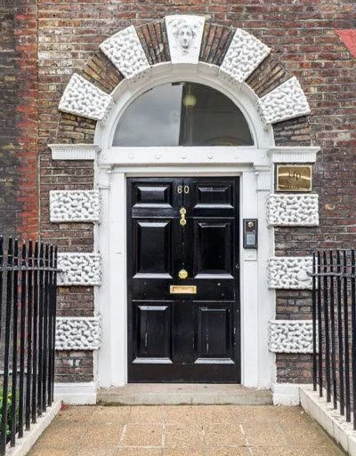 Black front door with the number 60, set in a brick building with an arched window above, surrounded by decorative white stone blocks, flanked by black metal railings.