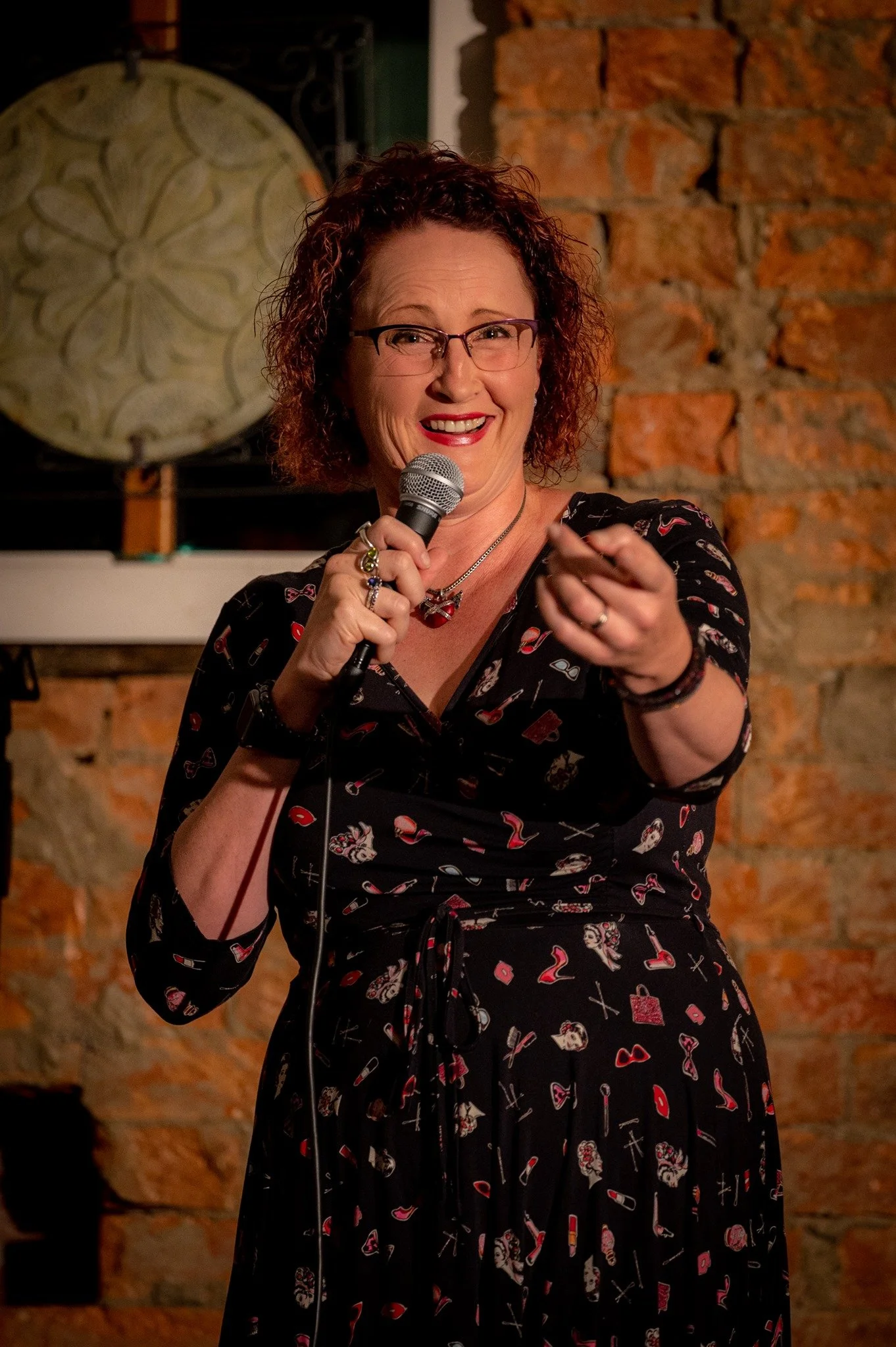 A woman with curly red hair and glasses smiling and pointing while holding a microphone on a stage with a brick wall background.