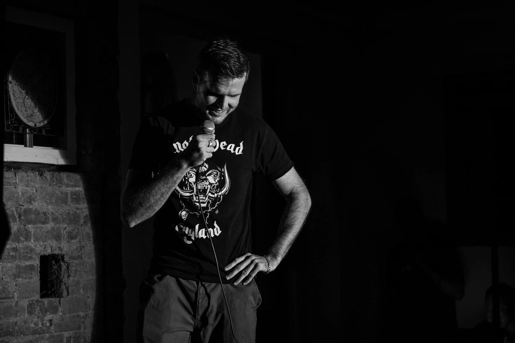 Black and white photo of a man standing indoors, holding a microphone, with a brick wall and a window in the background, wearing a black t-shirt with a graphic and the words 'Not Dead England'.